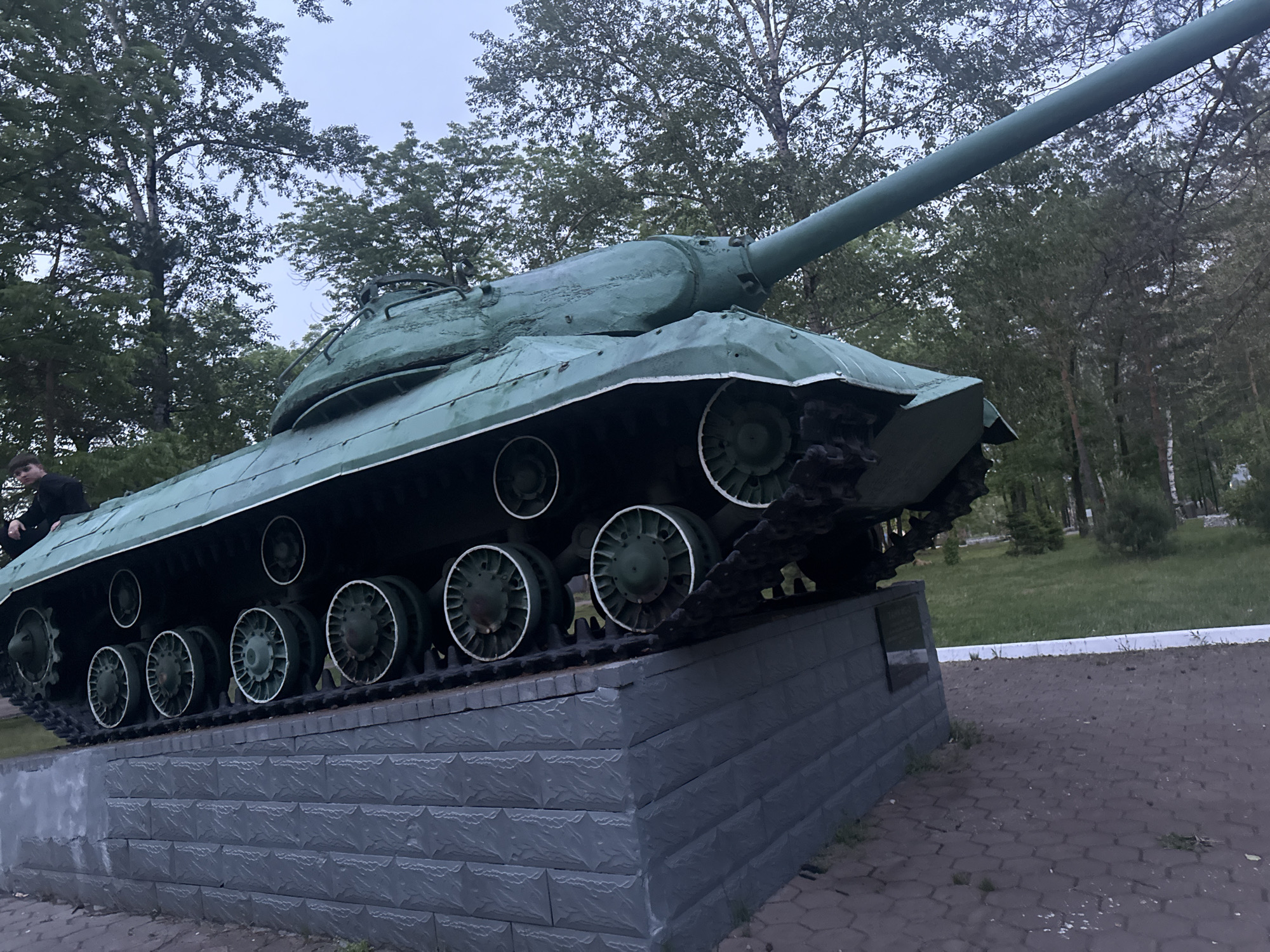The image displays a large, olive-green tank mounted on a grey stone pedestal in what appears to be a public park or square in Birobidzhan, Russia. The tank, identifiable by its distinct rounded turret, long gun barrel, and sloped hull, faces towards the right side of the frame, positioned at an upward angle on its display. Its dark tracks and road wheels with lighter rims are clearly visible along the side of the hull.
A person, dressed in dark attire, is partially visible on the far left, appearing to sit or lean against the tank's hull. The background is filled with lush green foliage from numerous deciduous and coniferous trees, suggesting an outdoor, natural setting. The sky above is overcast and light grey, indicating either a cloudy day or dusk.
The tank is supported by a robust pedestal made of grey, textured concrete blocks, designed with multiple layers. A dark, rectangular plaque is present on the side of the pedestal, though no specific text is legible. The ground around the monument is paved with interlocking stones, transitioning into grassy areas further back. The overall impression is that of a military monument or memorial within a landscaped public space.