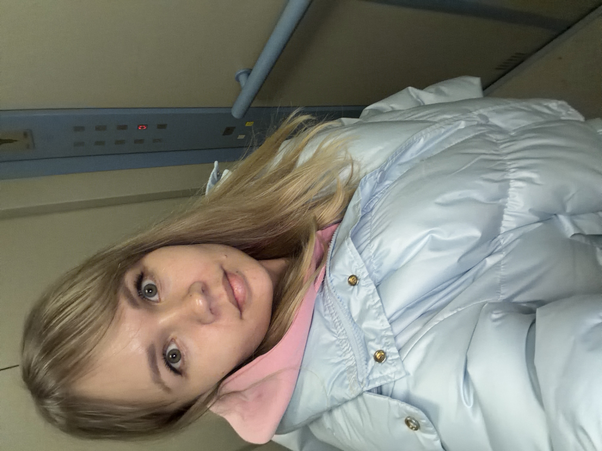 A young woman with long, blonde hair and light-colored eyes is featured in a close-up selfie, looking directly at the camera with a calm expression. She is wearing a light blue puffer jacket, fastened with gold-colored snap buttons, over a pink hoodie or top.
The setting appears to be an indoor, enclosed space, possibly a train carriage or an elevator. Behind her, a light-colored wall or ceiling provides the background. A distinctive blue panel, equipped with multiple square buttons and a centrally illuminated red display, is visible in the upper left area of the frame. The red display shows a symbol or digit, possibly "0". Above this panel, a silver-colored grab bar or handle runs horizontally. Next to the woman's head, there is a light-colored, possibly white, pillow or bunched fabric, suggesting she might be resting or lying down.
The lighting is soft and even, consistent with indoor artificial illumination. The image is captured from a slightly upward and rotated angle, with the woman's head positioned on the left side of the frame. Her attire suggests cooler weather. No other discernible text is visible in the image beyond the red display.