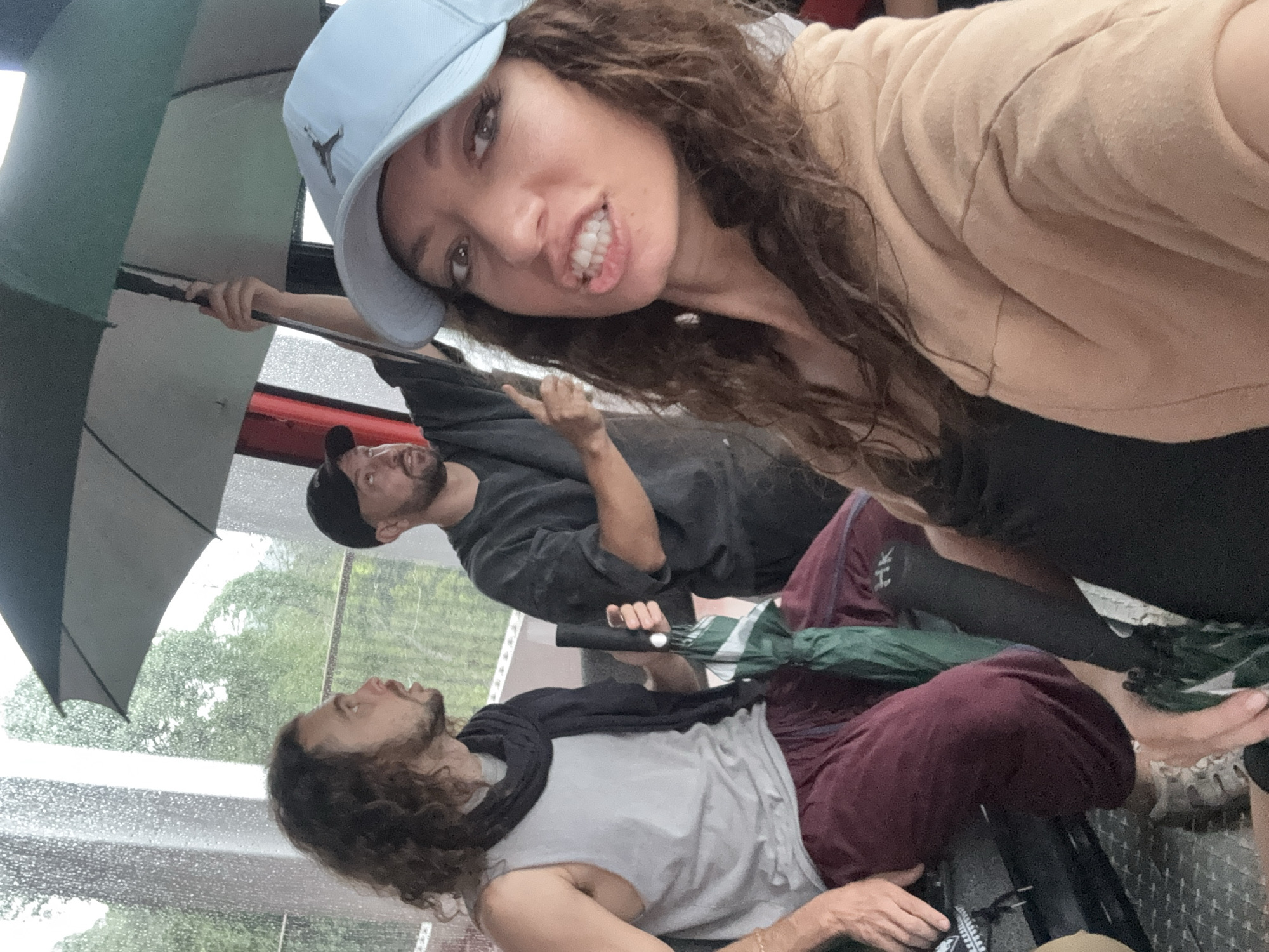 A group of three individuals is captured in a close-up, angled selfie, seemingly inside an enclosed space like a vehicle or cable car, amidst rainy weather in Đà Nẵng, Vietnam.
In the immediate foreground, a woman with long, curly brown hair wears a light blue baseball cap featuring a black Jordan (jumpman) logo. She looks directly at the camera with a playful, exaggerated expression, baring her teeth. She wears a light brown or tan jacket over a dark top.
Behind her and slightly to the left, a man in a dark cap and t-shirt holds a large black and grey umbrella. He looks upwards and to the side, away from the camera, with a slight beard visible. His right arm is raised, with his index finger pointing.
Further back, another man with long curly hair and a beard is reclined or lying down. He wears a light grey sleeveless t-shirt, a dark scarf around his neck, and maroon pants. A partially folded dark green and black umbrella rests beside him. His bare feet are visible, suggesting a relaxed posture.
The background shows rain streaks on a window, through which lush green trees and a blurred outdoor landscape are visible, indicating an overcast or rainy daytime. The floor appears to be a textured metal surface, possibly diamond plate. The presence of multiple umbrellas further emphasizes the rainy conditions outside. The overall mood conveyed by the woman's expression is lighthearted and candid.