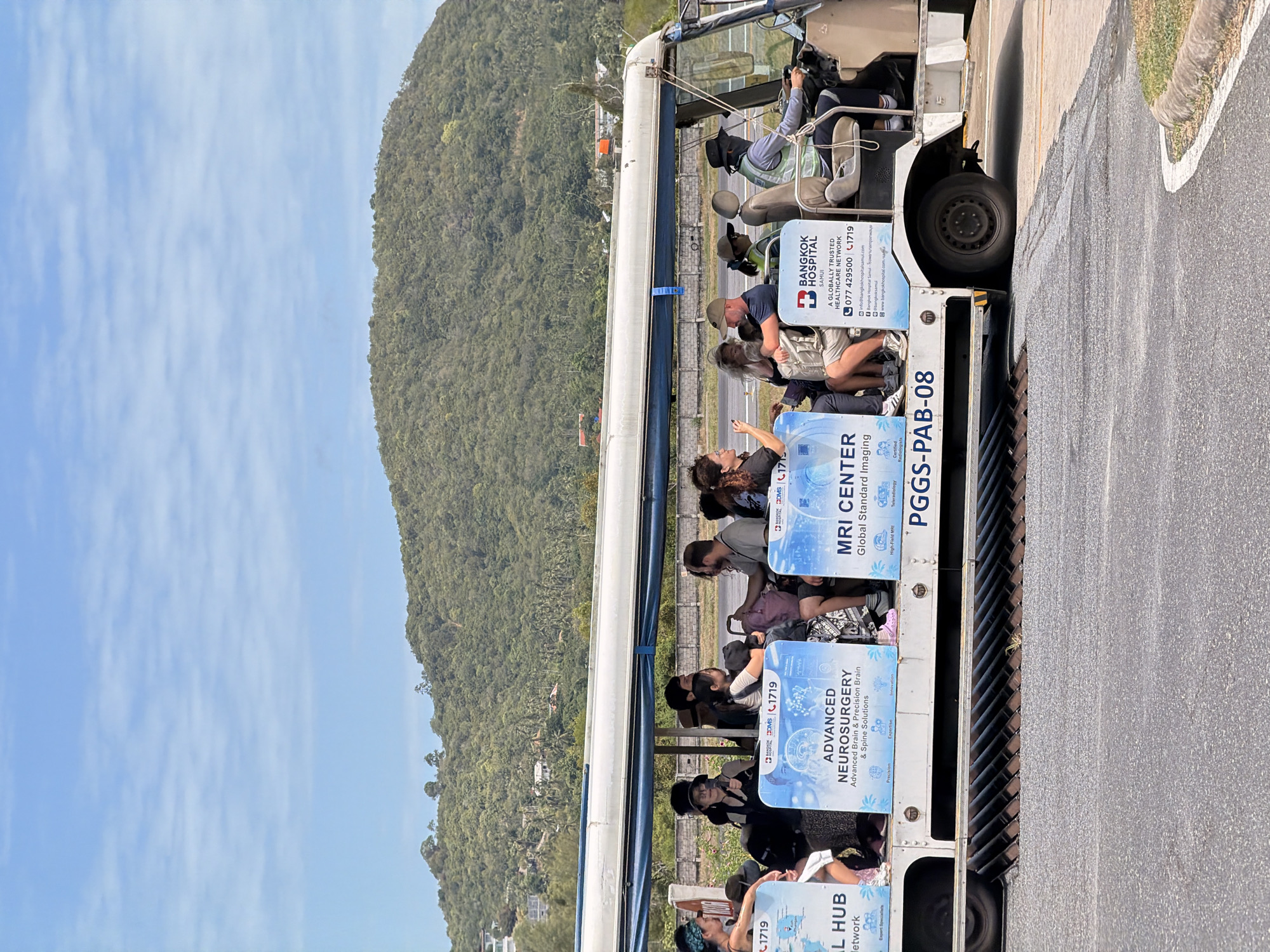 The image shows an open-air shuttle bus filled with passengers driving on a paved surface, likely an airport tarmac, in Ko Samui District, Thailand. The bus is white with blue trim and displays advertisements for Bangkok Hospital Samui, highlighting services such as "Advanced Neurosurgery" and "MRI Center." A lush, green, tree-covered mountain rises in the background under a partly cloudy sky. The passengers, a mix of men and women, are seated and appear to be on a tour or transfer. Some are looking at their phones, others are conversing, and one person is driving the vehicle. The overall scene suggests a typical tourist transportation experience in a tropical location.