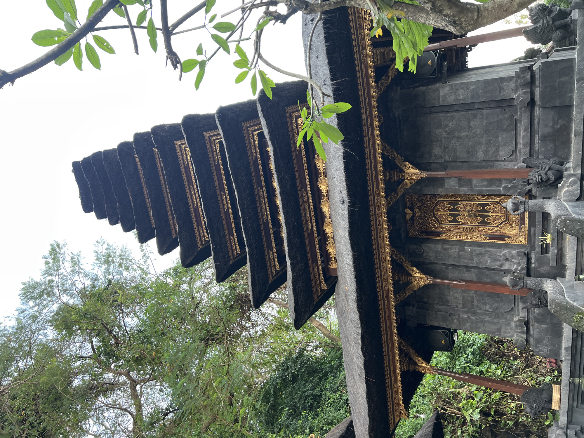 This image captures a tiered temple structure in Dawan, Indonesia. The temple features dark, stone walls and multiple layers of thatched roofs, each adorned with intricate golden trim. A large, ornate golden door is visible on the front of the structure. The temple is surrounded by lush green foliage and trees. The sky is overcast. Small statues of mythical creatures flank the entrance.