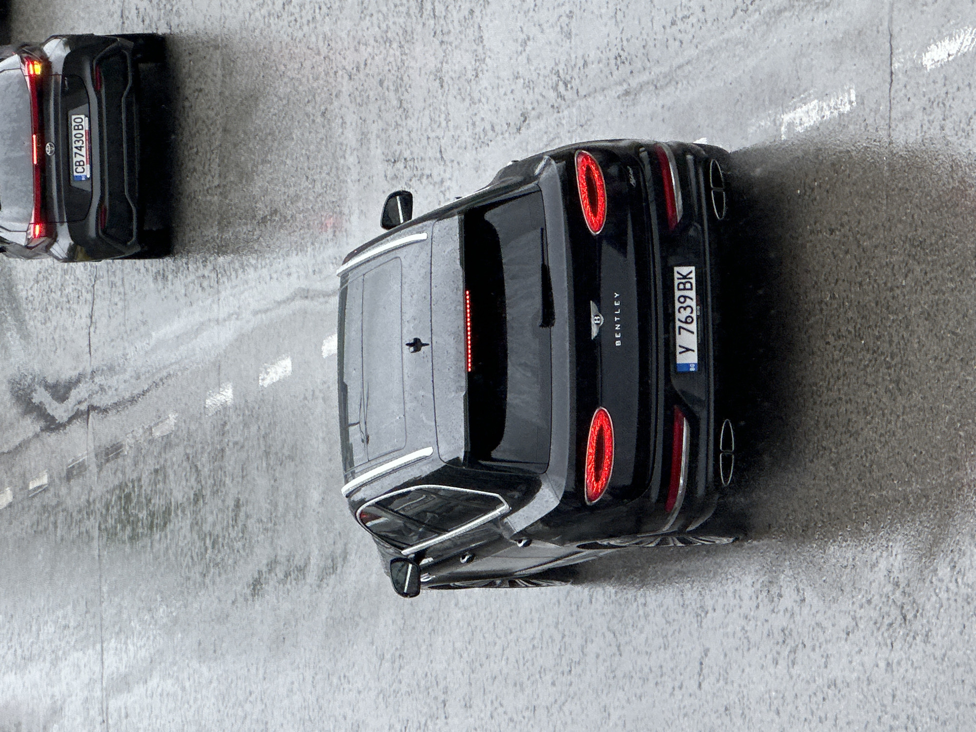 The image shows two cars on a wet road, likely in Veliko Tarnovo, Bulgaria. The car in the foreground is a black Bentley with license plate number "Y 7639 BK." The other car in the image is a smaller black vehicle, partially visible in the upper left corner, with a license plate number that appears to be "CB 7430 BO." The road is marked with white lane dividers, and the overall scene suggests wet or rainy weather conditions.