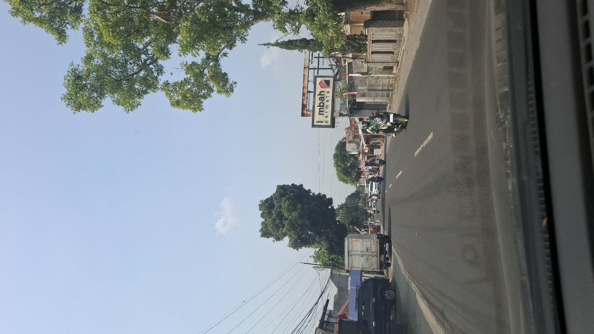 A perspective shot from inside a vehicle shows a road in Warungkondang, Indonesia, on a clear, sunny day. The road is paved and has white lane markings. Several vehicles, including cars and motorcycles, are traveling on the road. On the left side of the road, there is a black SUV and a white truck. On the right side, there are buildings, fences, and trees. A large sign with the text "I mbah permata" is visible on the right. The sky is blue with a few wispy clouds. The overall scene depicts a typical day on a road in a town.