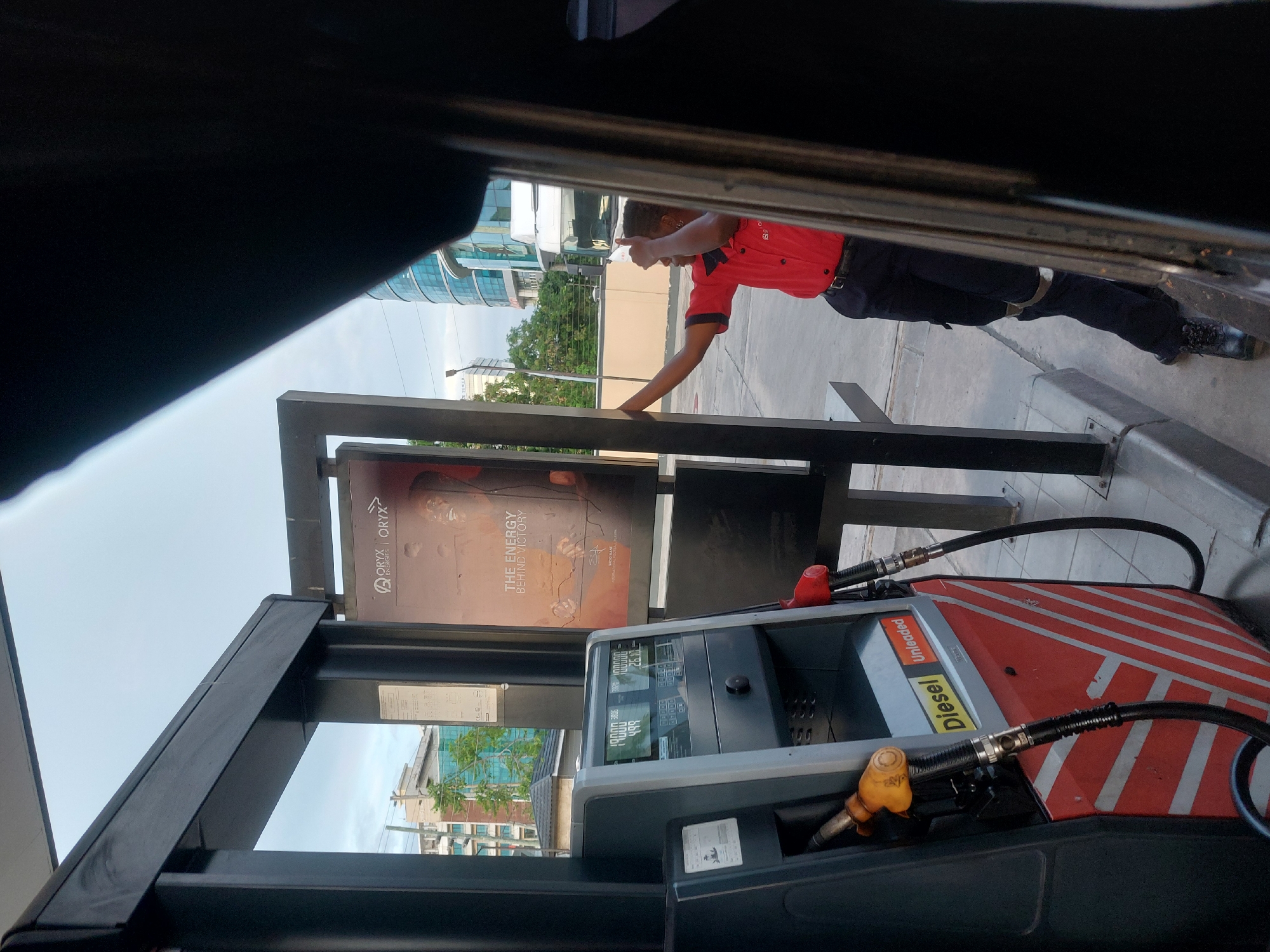 A gas station attendant, wearing a red uniform shirt and black pants, leans against a black metal frame holding an advertisement sign at an Oryx gas station. The sign reads "Oryx Energies: The Energy Behind Victory". A gas pump with a red and white striped design and labeled "Diesel Unleaded" is in the foreground. The view is from inside a vehicle. The sky is overcast. The location is likely in Dar es-Salaam, Tanzania, based on the Oryx gas station branding. A modern building is visible in the background.