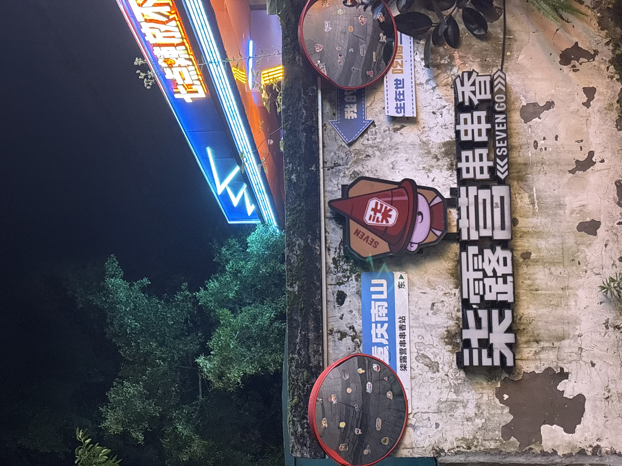 The image depicts a narrow vertical view of a weathered, moss-covered wall in the Nan'an District of China, likely outdoors, at night. The wall is adorned with various signs and decorations. A large, bright, neon sign with Chinese characters is partially visible on the left side, oriented diagonally.
Attached to the wall are two convex circular mirrors with red rims. The mirrors feature numerous small stickers or decals. Between the mirrors is a blue sign with white Chinese characters stating "慶南山" (Qingnan Mountain), underneath which is a smaller line of text in Chinese. There is an arrow and a sign which states "生在世" (born in this world). There is a cartoon character icon of a person wearing a red hat.
To the right, a vertical sign with Chinese characters and the words "SEVEN GO" are mounted to the wall. The wall itself is discolored and peeling. The left side of the image features foliage and dark sky.