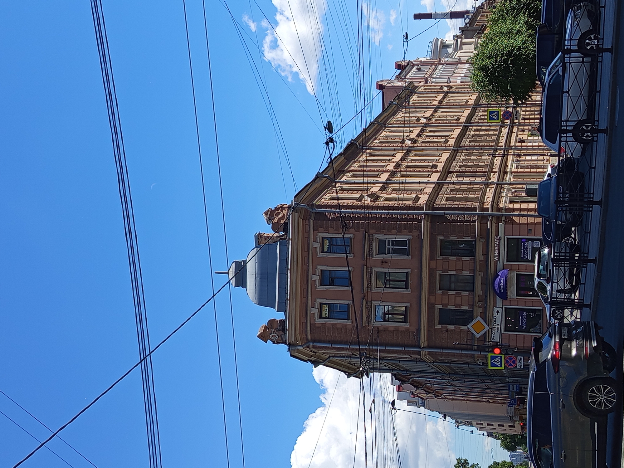 The image depicts a street scene in Saint Petersburg, Russia. A prominent, angular building with a domed roof and ornate architectural details dominates the center of the frame. The building is painted in shades of peach and ochre, with numerous windows. Multiple signs are visible on the building, including "WineState" and "FOR SALE" signs in English. A traffic light displays a red signal, and a pedestrian crossing sign is present. Several cars, including a dark SUV in the foreground, are stopped at the intersection. The sky is clear blue with scattered white clouds, and a crescent moon is faintly visible. Power lines crisscross the sky above the buildings and street. The environment suggests an urban setting in the daytime.
