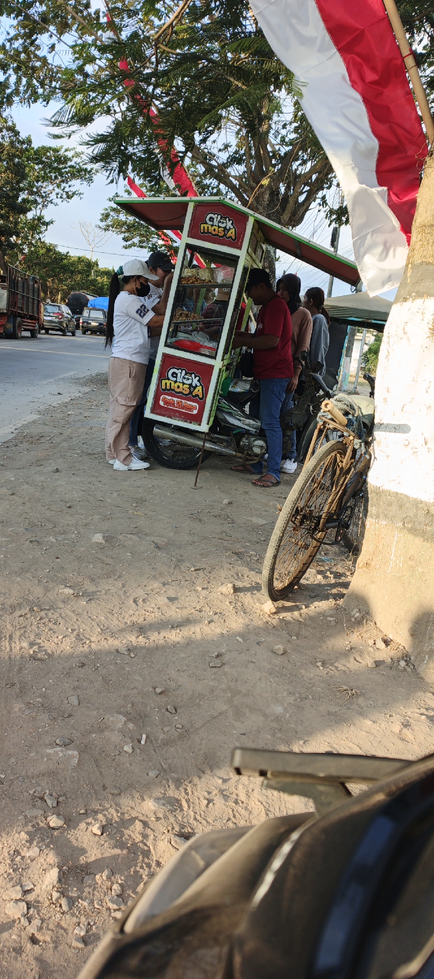 A group of people are gathered around a food cart, which is mounted on a motorcycle. The food cart is green and red and displays the text "Cilok Mas A". It appears to be selling snacks. The scene takes place on the side of a road, with cars and a truck visible in the background. Trees and Indonesian flags are also present. The ground is dirt and gravel. The overall impression is that of a busy street food vendor.