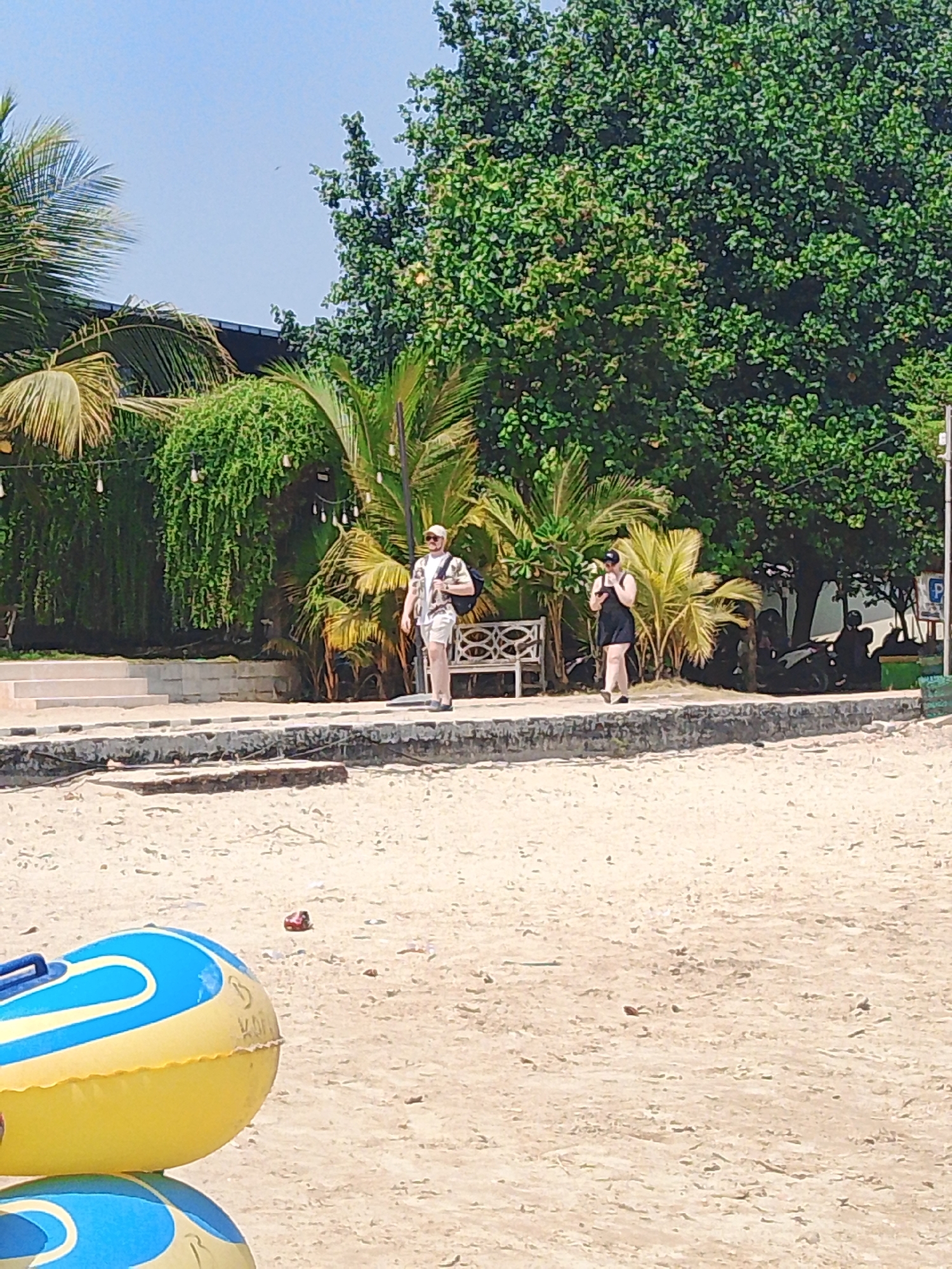 The image depicts a beach scene in Tegalsambi, Indonesia. In the foreground, a portion of a yellow and blue inflatable tube rests on the sand. The midground shows a paved walkway bordering a sandy area, with lush green trees and palm trees forming a backdrop. Two individuals are visible walking on the walkway. A man, wearing a patterned shirt, shorts, sunglasses, and a hat, walks towards the left. Behind him, a woman in a dark dress and a cap is also walking towards the left. A white bench is situated on the walkway between the two individuals. In the background, several motorcycles are parked near some trees. The sky is clear and blue.
