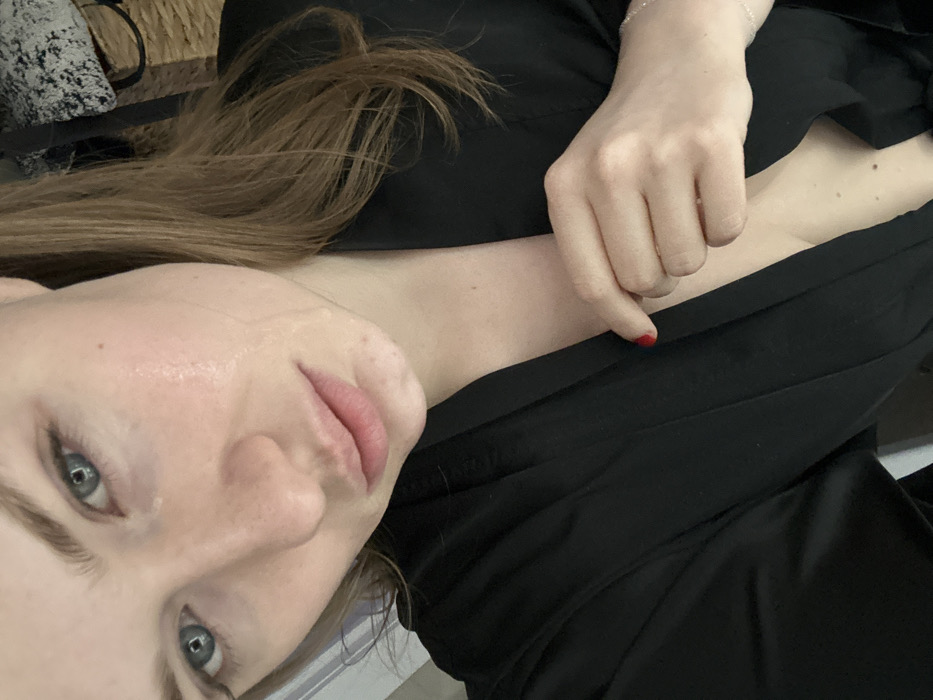 A woman angles her face towards the camera, showcasing light eyes, fair skin, and light brown hair. She is wearing a black garment. Her hand rests on the fabric, with a single fingernail painted red. The image is cropped, primarily focusing on her face and upper body. The background is somewhat blurred, featuring muted tones and shapes.