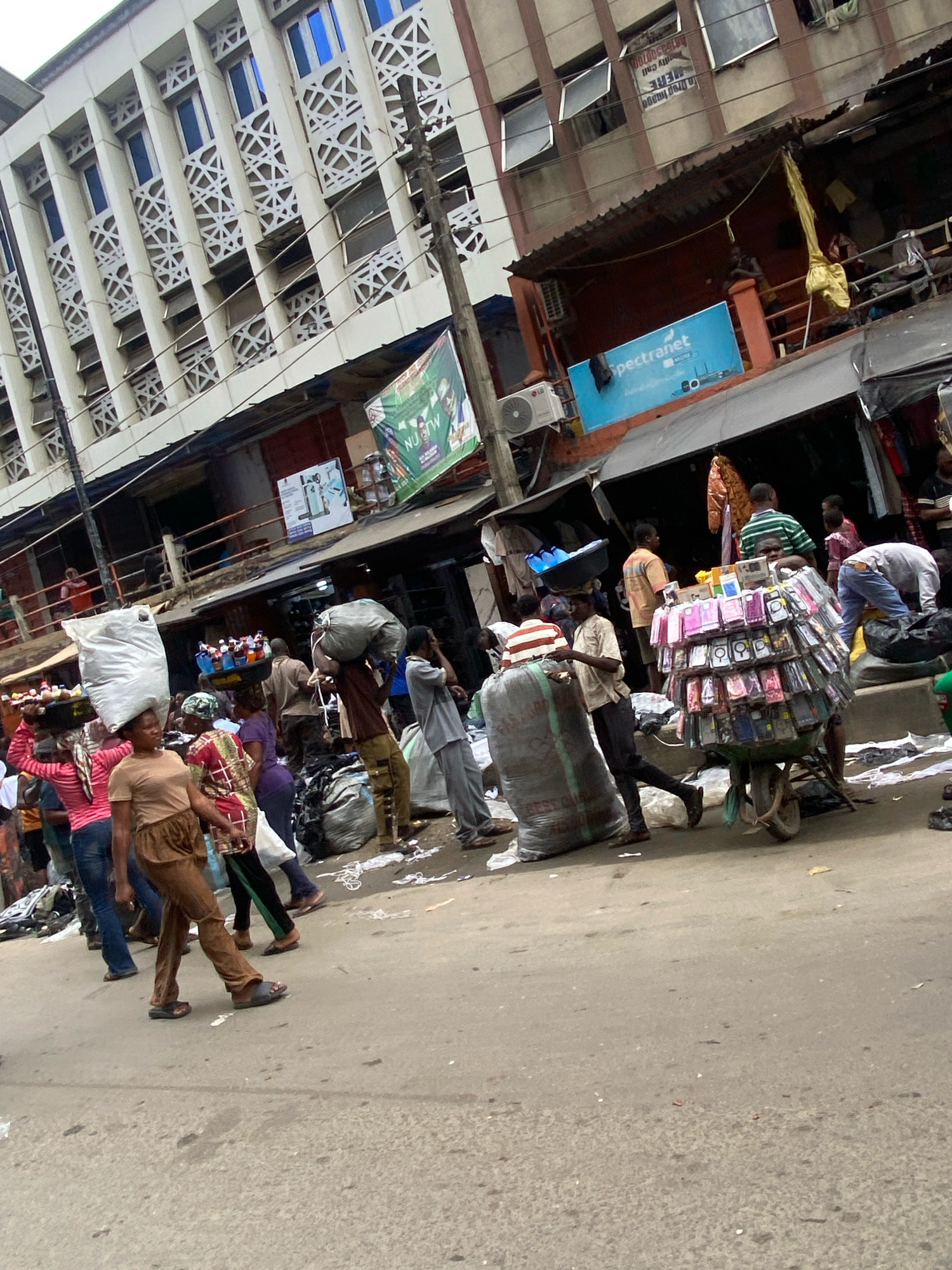 This image captures a bustling street scene on Lagos Island, Nigeria. A long, multi-story building with a distinctive patterned facade lines the left side of the street. Numerous people are engaged in commerce and daily activities. In the foreground, a woman in a pink top and brown skirt carries a large white bag on her head, walking alongside another woman in a brown top and skirt. To the right, a man pushes a cart laden with numerous phone cases. Several other individuals are seen carrying goods, interacting, or simply moving through the crowded street. Bags of various sizes and materials, as well as bottles of liquids, are visible. The environment appears to be an outdoor market or a busy commercial area with awnings and stalls.