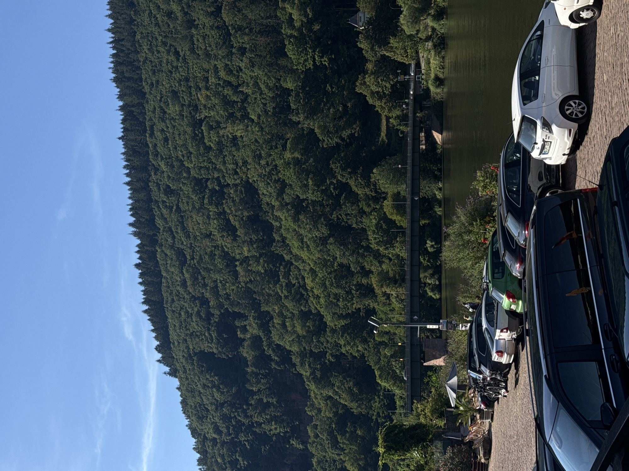 In Neckargemünd, Germany, a sunny day is depicted with a river flowing through a town nestled at the base of a verdant, tree-covered hill. The riverbank is lined with parked cars, including a silver hatchback and a white hatchback parked side-by-side, with a green hatchback positioned behind them. A pedestrian bridge spans the river in the background. The hill is densely forested, with a clear blue sky overhead. The scene suggests a peaceful day in a picturesque German town.