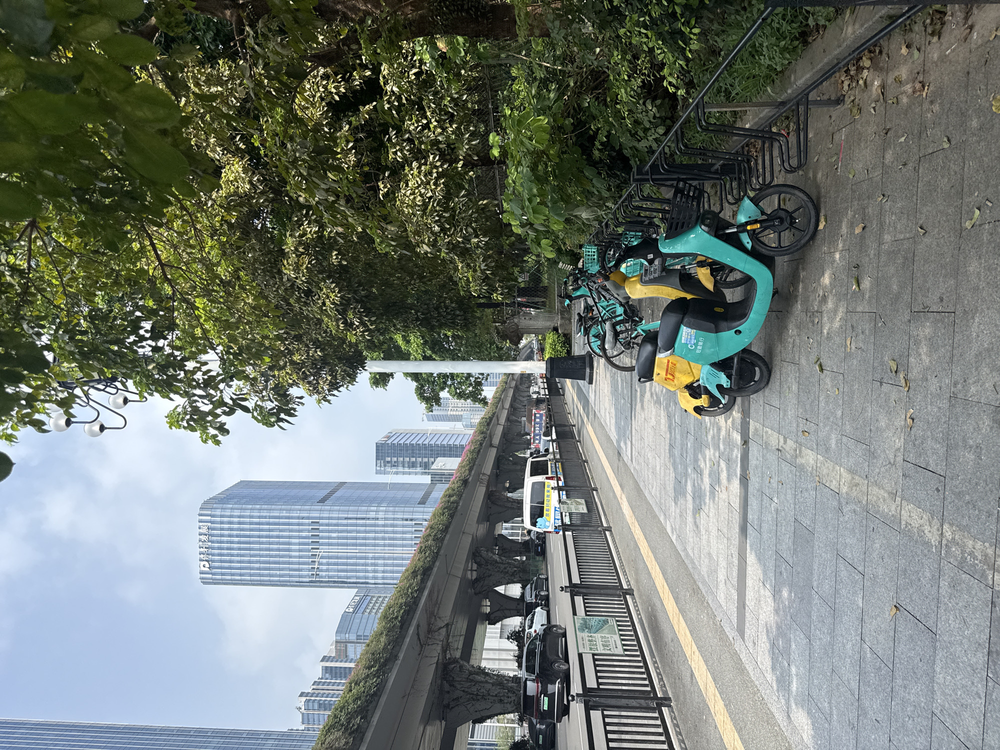 The image depicts a street scene in Huangpu District, China. In the foreground, a sidewalk is paved with gray tiles, with a yellow tactile paving strip along the edge. Several shared electric scooters, colored turquoise and yellow, are parked in a rack. Beyond the sidewalk, a black metal fence separates the pedestrian area from a multi-lane road. Cars and a white van are visible on the road. Overhead, an elevated structure, possibly a highway or metro line, is supported by large, vine-covered concrete pillars. Tall modern buildings with glass facades rise in the background, and lush green trees line the right side of the image, casting shadows on the sidewalk. The sky is partly cloudy.