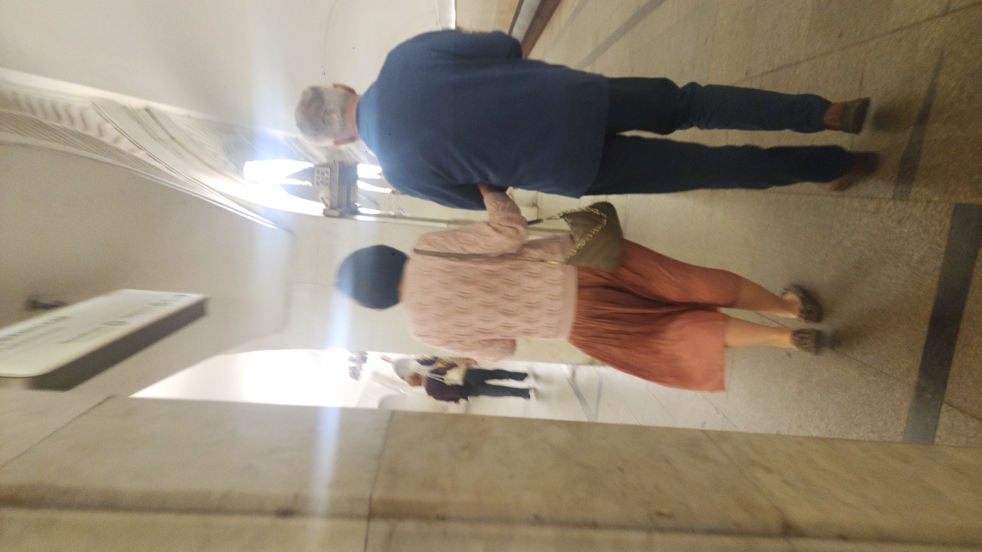 An eye-level, blurred, slightly out-of-focus shot depicts two people walking in what appears to be an underground subway station or tunnel in Unknown, Unknown. The foreground features part of the architecture of the station, showing what appears to be a marble or stone wall on the left side. A sign in a white rectangular case is mounted on the wall. The background shows people walking in a hallway. The lighting is strong, creating glare in the image. In the center foreground, a man and a woman walk away from the camera. The man is on the left side of the frame and is wearing a dark blue sweater, dark-colored pants, and brown shoes. The woman, located slightly behind the man, is wearing a light pink cardigan with a brown purse hanging from her shoulder, an orange pleated skirt, a dark cap, and patterned shoes.