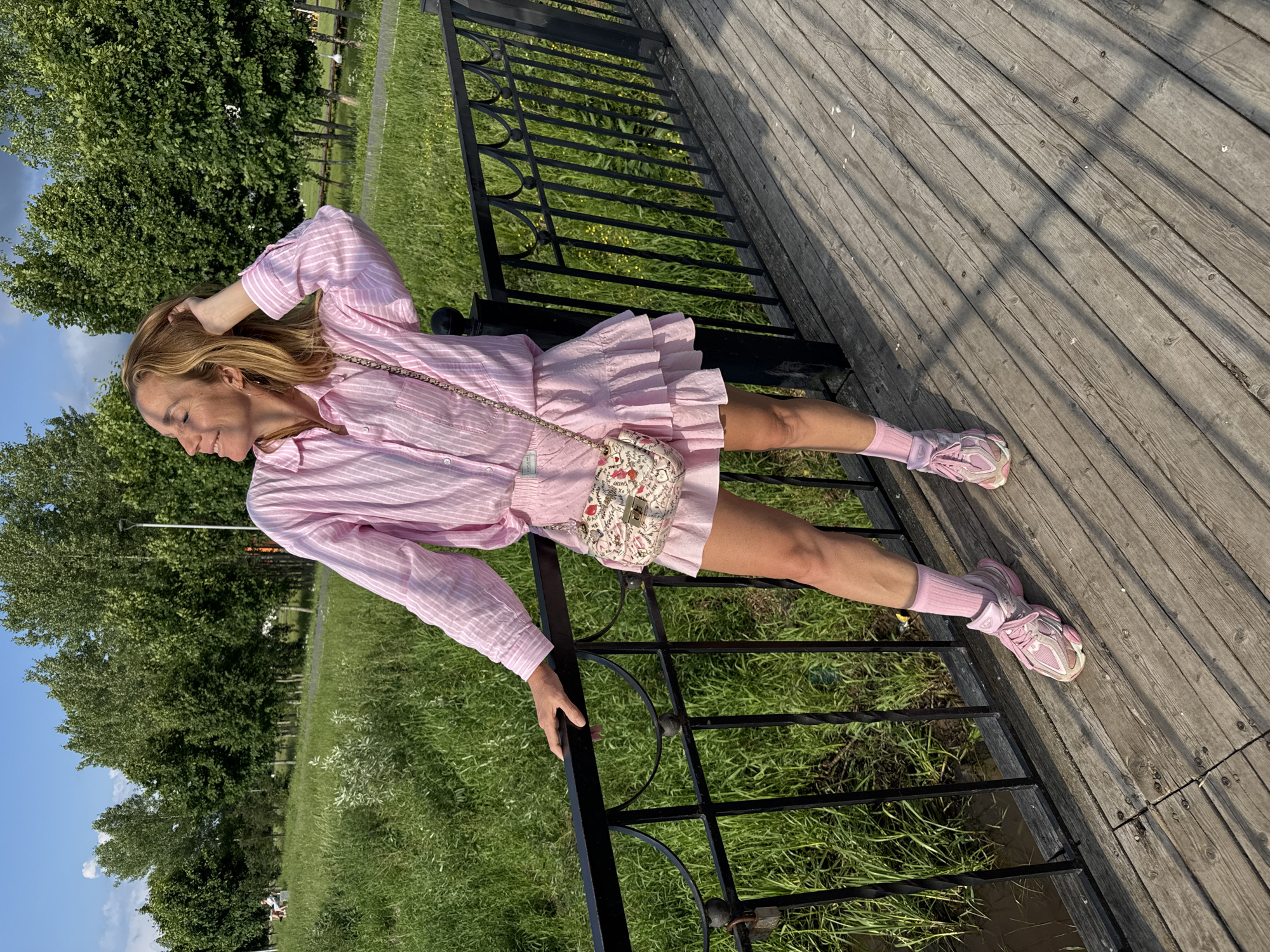 A woman is standing on a wooden walkway next to a black metal fence, leaning with her left hand on the fence. She is wearing a pink striped button-down shirt, a pink ruffled skirt, pink socks, and pink sneakers. A small patterned purse is hanging from her left shoulder. Her right hand is in her hair, and she is looking to her right with a slight smile. The background shows a grassy area with trees and a blue sky with some clouds. The scene is located in a park in the city of Kirishi, Russia.