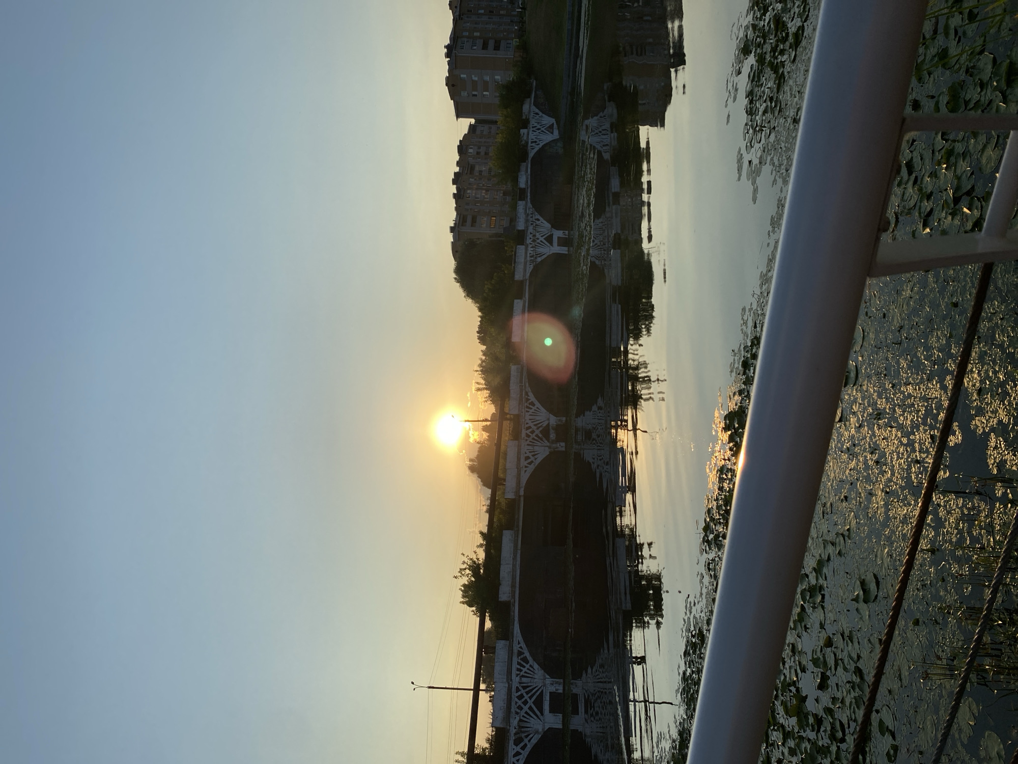 The image depicts a sunset over a body of water in Votkinsk, Russia. In the background, a white arched bridge spans the water, with apartment buildings and trees visible on the far bank. The setting sun casts a warm glow across the sky and reflects on the water, which is partially covered with lily pads. In the foreground, a metal railing and rope barrier are visible, suggesting a waterfront promenade or park. The scene is tranquil, with no people or animals present. The overall atmosphere is serene and picturesque, capturing the beauty of a summer evening in Votkinsk.