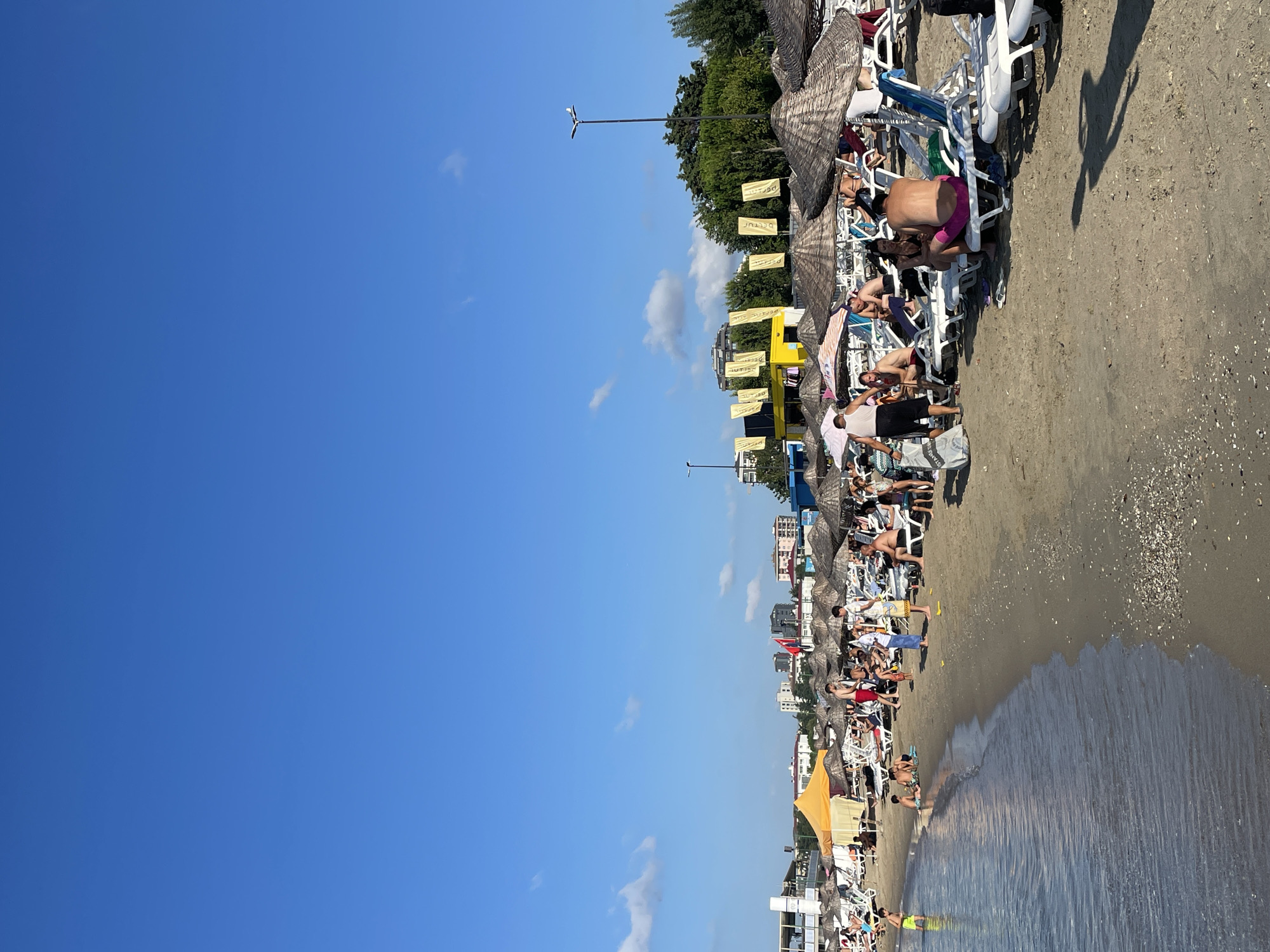 The image depicts a crowded beach scene on a sunny day in Kadıköy, Turkey. The foreground shows the gentle waves of the sea lapping against a sandy shore littered with shells. The beach is lined with numerous rows of beach chairs and umbrellas, creating a lively atmosphere.
A variety of people are scattered across the beach, some lounging on chairs, others standing or walking. Several individuals are visible in the shallow water near the shore, including a young boy in yellow shorts. In the mid-ground, a yellow tent provides shade, and behind it, a row of buildings and trees form the backdrop.
Further back, more umbrellas and beach chairs are visible, with a collection of people enjoying the beach. A bright yellow structure with flags is prominent, and a tall lamppost with a light fixture is also present. The sky is a clear, vibrant blue with a few scattered white clouds.
The scene suggests a typical summer day at a popular beach resort, with ample recreational activity and relaxation.