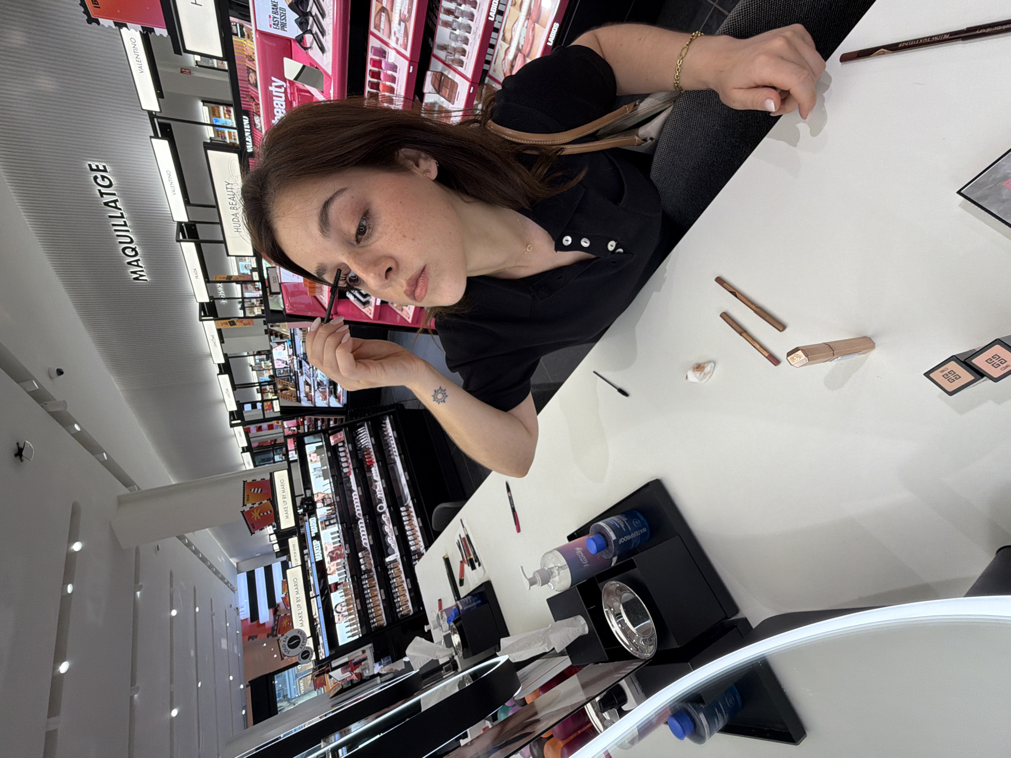 In a brightly lit cosmetics store, identified by a sign reading "MAQUILLATGE" and product displays for brands like "HUDA BEAUTY" and "VALENTINO," a young woman with brown hair is applying mascara. She is seated at a white counter, facing a mirror that reflects her, with various makeup products scattered on the surface. These include lipstick, eyeliner, and what appear to be small makeup palettes. A bottle of hand sanitizer and another bottle labeled "WATERPROOF" are also visible. The woman is wearing a black collared shirt and has a small tattoo of a sun on her left arm. The background reveals rows of makeup products and a digital display promoting "MAKE UP BY MARIO." The overall scene suggests a beauty retail environment, potentially in Barcelona, Spain, given the Catalan word "MAQUILLATGE" for makeup.