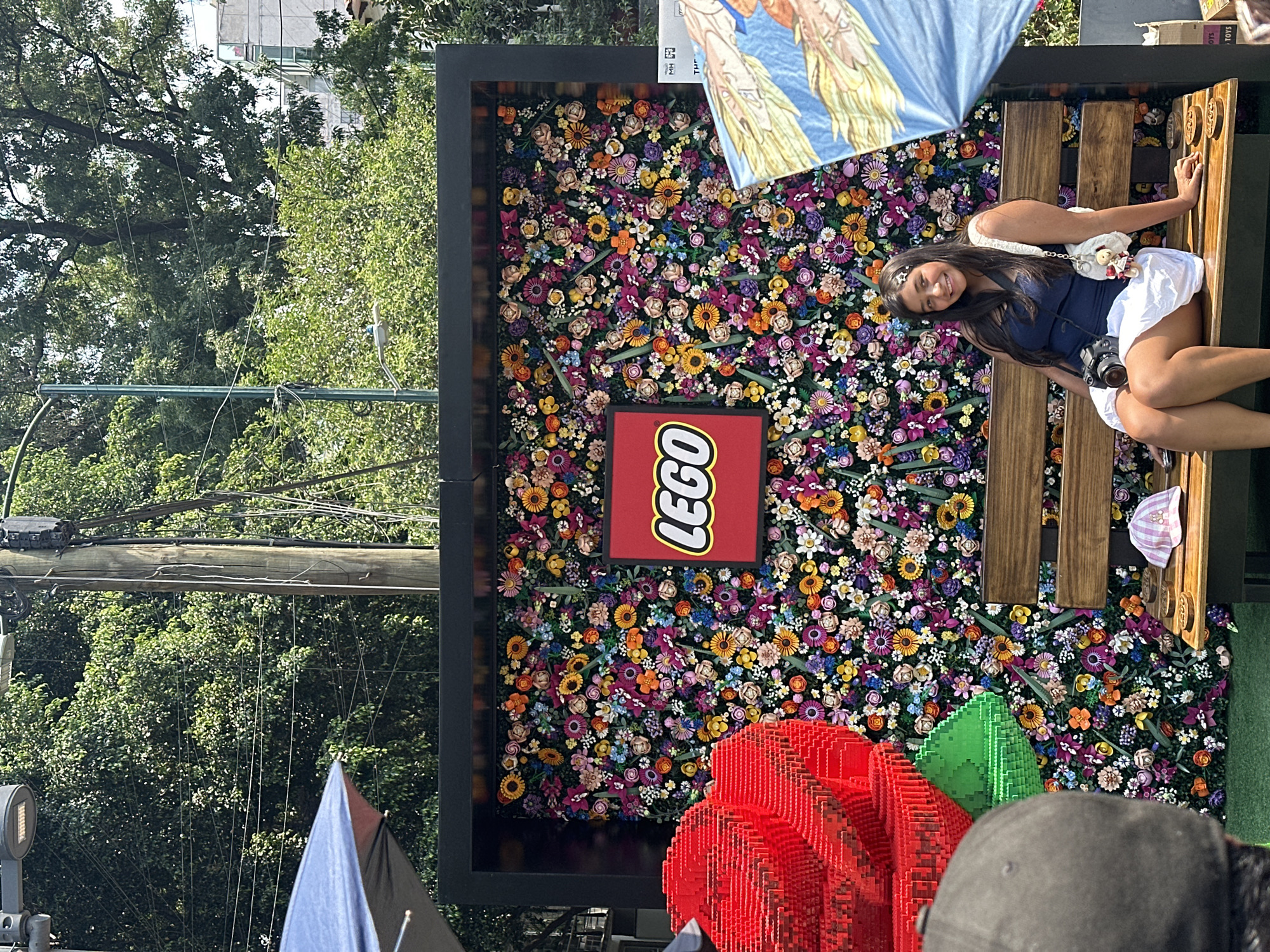 A medium shot shows a young woman sitting on a wooden bench in front of a flower wall with a Lego logo in Mexico City, Mexico. The flower wall is a backdrop of colorful flowers, including sunflowers, in various shades of yellow, orange, and purple. In the lower-left corner of the image are two Lego sculptures, a red heart and a green triangle. A man's head and shoulder are visible in the lower-right corner. Behind the wall and woman are trees, wires, and streetlights. An advertisement is in the top-right corner, angled. The image appears to be taken outside during daylight. The woman is smiling at the camera. She has dark hair and is wearing a blue top and white skirt. She also has a camera on her lap.