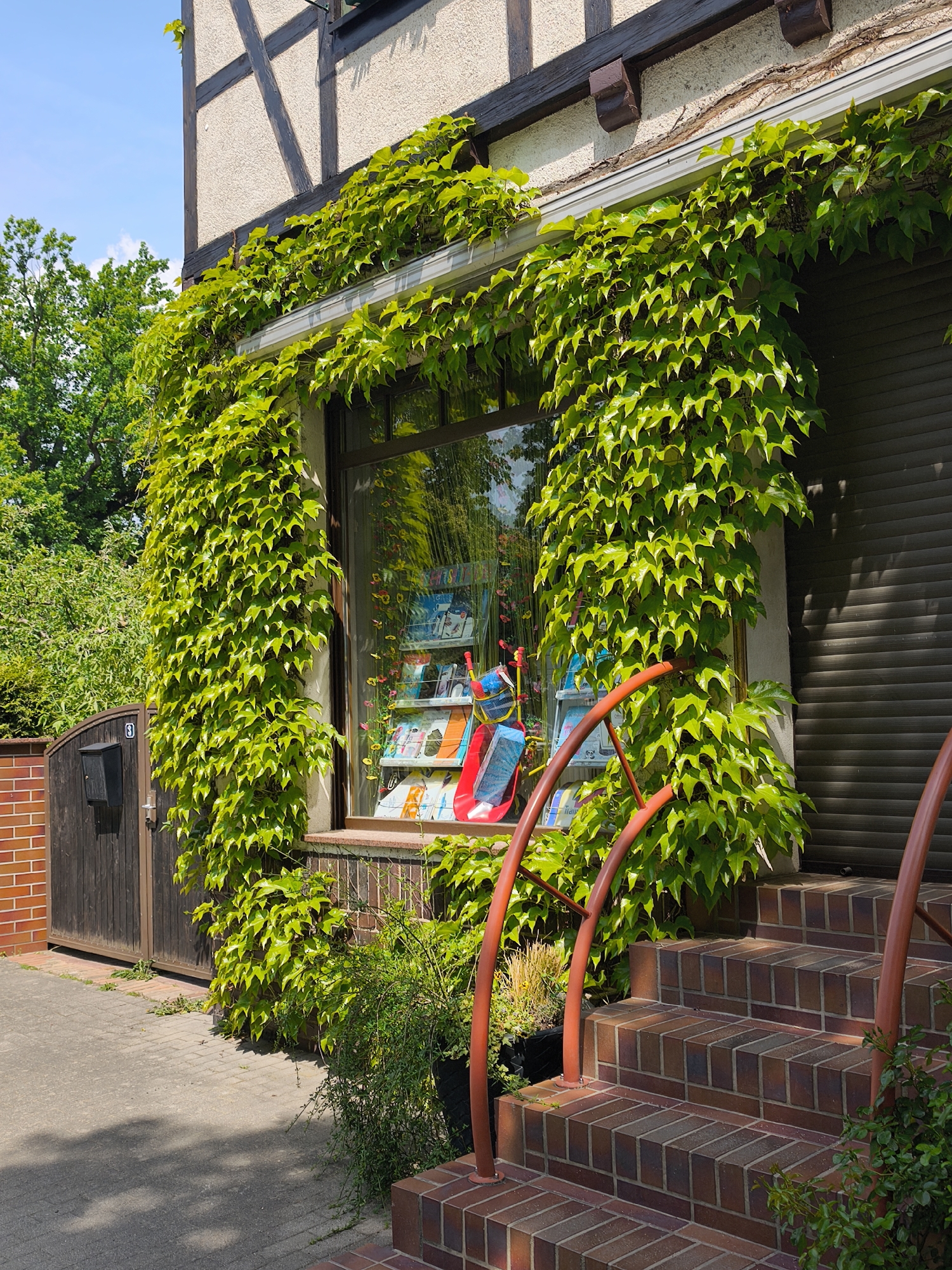 The image depicts the exterior of a building. The facade features a window and a shuttered section, both heavily overgrown with ivy. To the right, brick steps lead up to the building, with orange railings flanking the stairs. In front of the window are plants in pots, and the window itself displays items on shelves. A portion of a fence and gate are visible to the left of the building. The building's upper section is of a timber frame design. The location appears to be in a city.