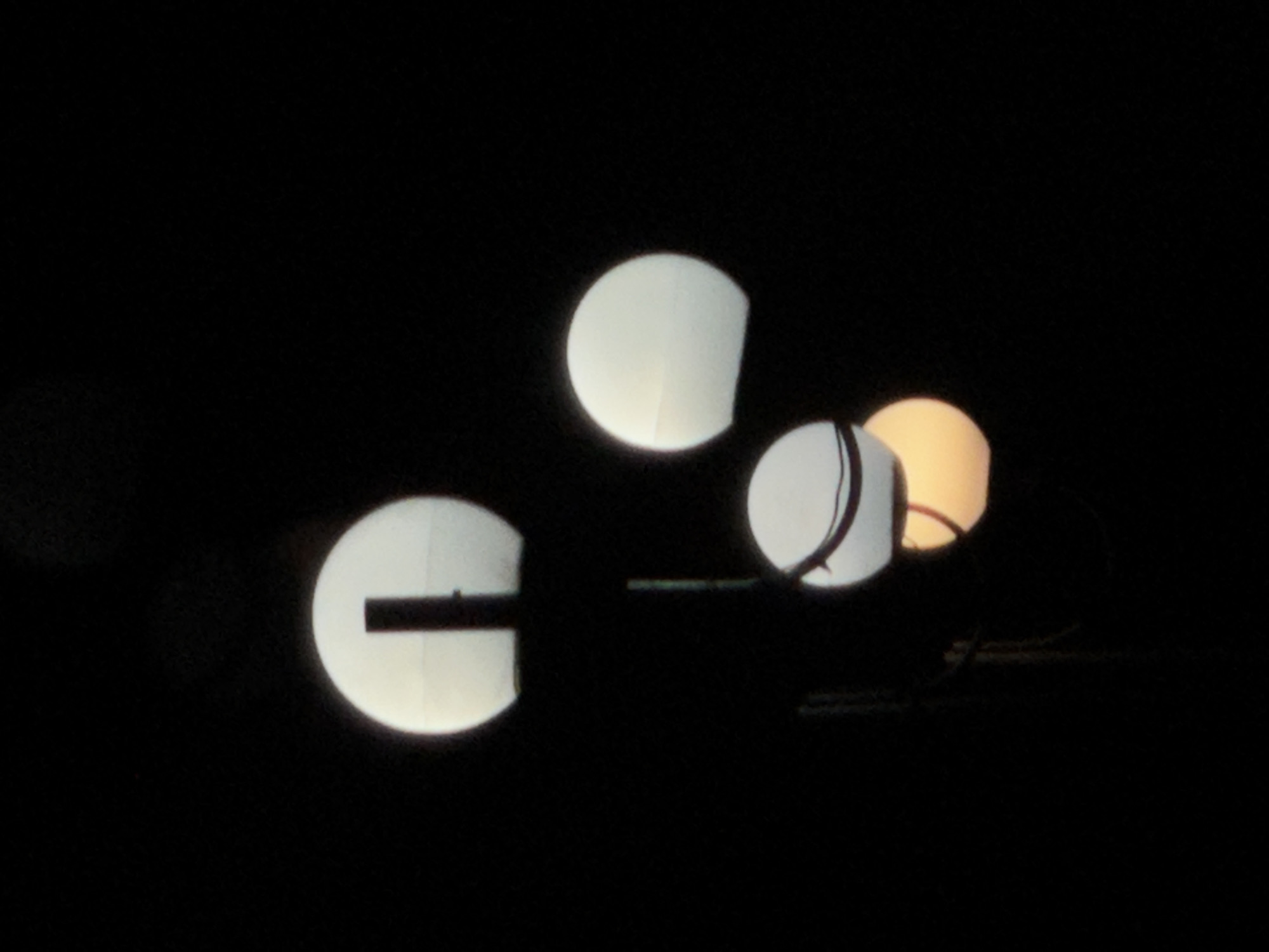 Three lanterns are visible in the darkness. One appears white with a horizontal black line bisecting it. Another is white with a thin black line wrapping around its side. The third lantern has a yellowish tint. Some wires or cables are present in the lower-right portion of the image. It is nighttime. The image location is Lehchevo, Bulgaria.