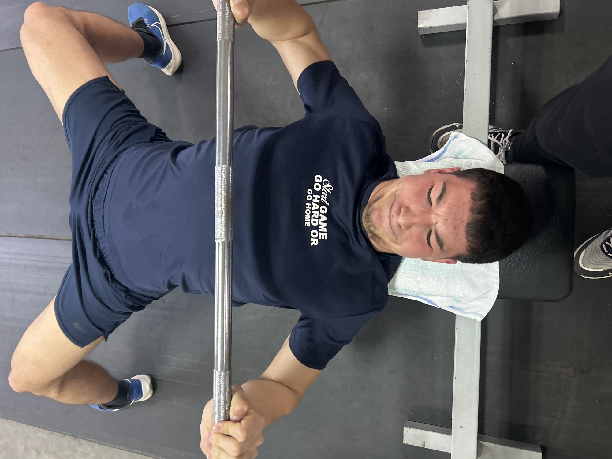 An overhead shot captures a young man performing a bench press exercise in what appears to be a gym in Zlatograd, Bulgaria. He is lying on a black weight bench, his head resting on a white and blue striped towel. His face is contorted in an expression of intense strain, indicating significant effort in lifting the barbell held in both hands above his chest.
The man is wearing a dark blue athletic t-shirt with short sleeves and matching dark blue athletic shorts. A white logo and text are visible on his shirt, reading "Start GAME GO HARD GO HOME". On his feet, he has blue and white athletic shoes on his left foot and a black and white athletic shoe on his right, the latter partially obscured by a black pant leg or shoe of another person, possibly a spotter. His legs are bent at the knees, with his feet flat on the dark, possibly rubberized, gym floor.
The shiny metal barbell, featuring knurled grips, is prominent across the center of the frame. To the right of the man's head, a grey metal support structure of the weight rack is visible, along with another person's shoe and leg. The overall setting is simple, focusing on the exercise and the man's exertion. The lighting suggests an indoor environment.
