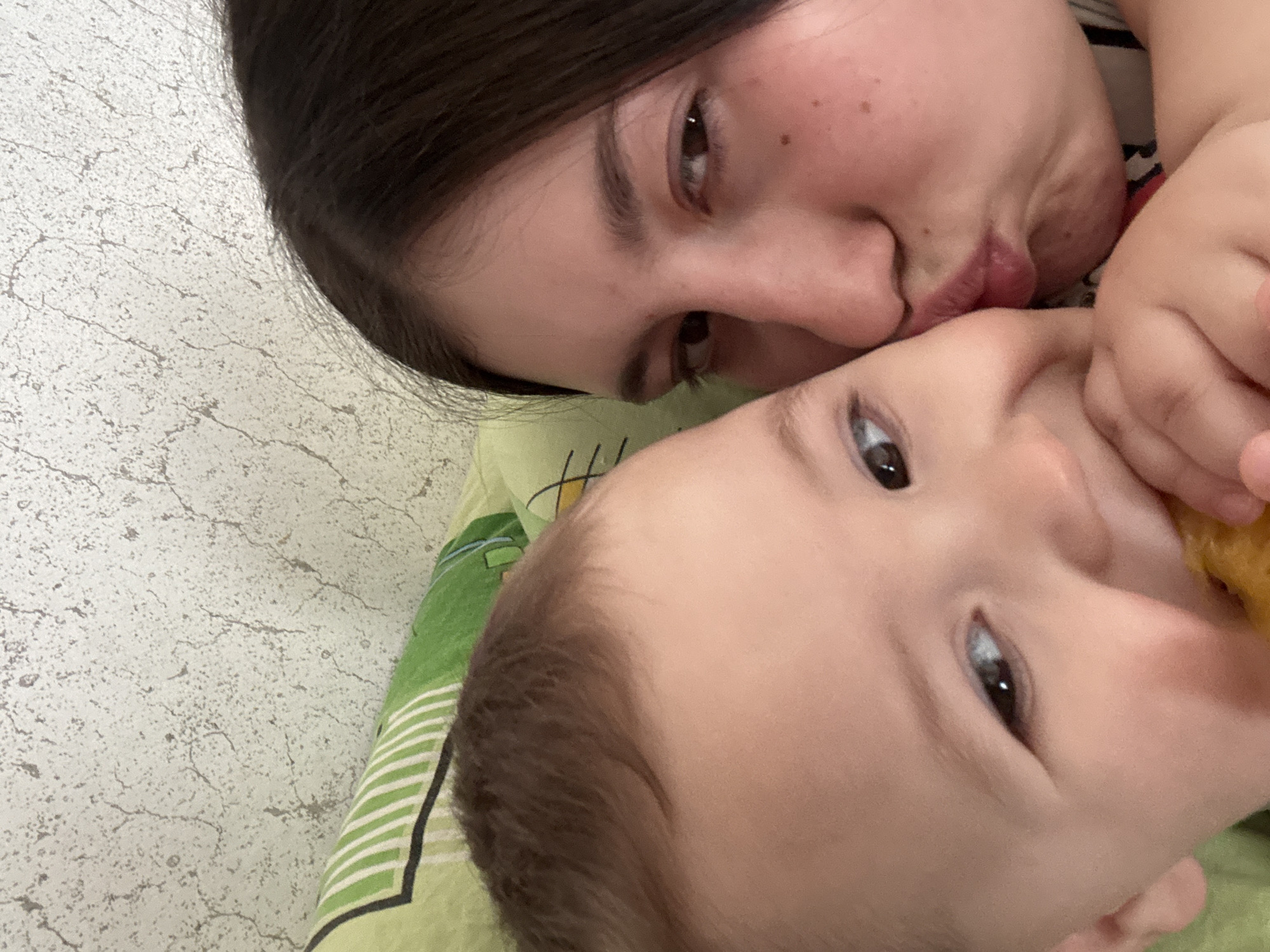 A woman and a baby are close together in an indoor setting. The woman has dark hair and is puckering her lips as if to kiss the baby. The baby has fair skin, dark hair, and is looking at the camera. The scene appears to be indoors, possibly on a bed, indicated by the green and white striped fabric. The location is Фоки, Russia.