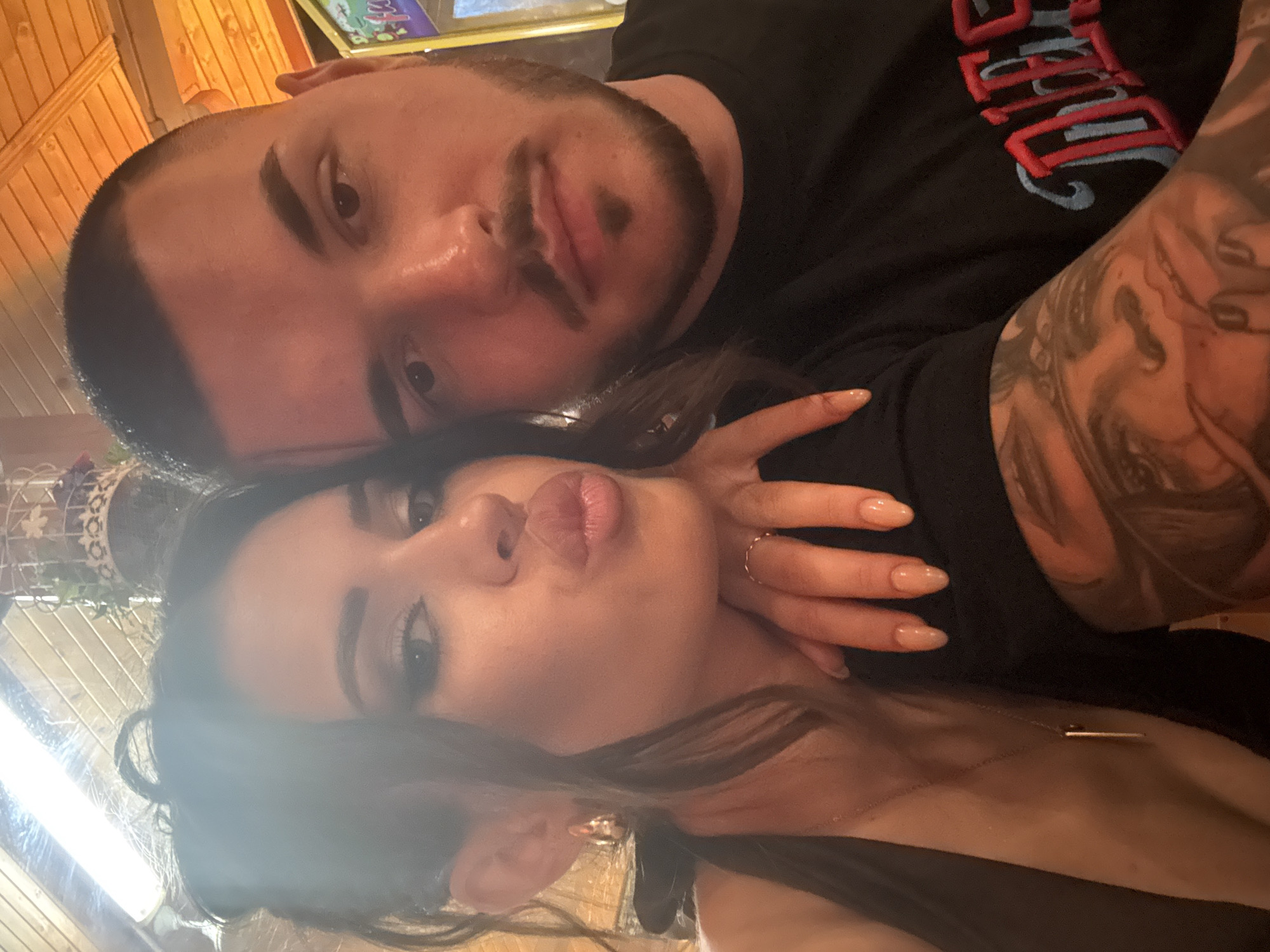 A forensic examination of the image reveals two individuals, a female and a male, captured in a close-up, selfie-style photograph within an indoor establishment in Debrashtitsa, Bulgaria.
The female, positioned on the left and closer to the camera, is an adult with dark hair, styled with strands framing her face. She wears full facial makeup, including defined eyebrows, dark eyeliner, and eyeshadow. Her lips are pursed in a "kiss face" expression, and her gaze is directed towards the camera. She is adorned with gold-toned hoop earrings, a thin gold-toned necklace featuring a bar pendant, and a thin gold-toned ring on her left ring finger. Her fingernails are long, oval-shaped, and appear to have a light-colored or clear polish. She is wearing a dark, possibly black, halter or tank-style top. Her right hand is visible, resting on the male's left shoulder/upper arm area.
The male, positioned to the right and slightly behind the female, is an adult with a short, buzzed dark haircut. He has a neatly trimmed goatee and mustache, with a short beard along his jawline. His expression is neutral, and his gaze is directed towards the camera. He is wearing a black t-shirt with partially visible embroidery on the right chest area, featuring red and blue lettering that appears to spell "DIES." His left arm is prominently displayed, covered by a detailed tattoo sleeve depicting a stylized face, possibly a woman with a bandana, among other patterns.
The background indicates an indoor setting with warm, artificial lighting. A long, bright light fixture is visible on a wooden-paneled ceiling at the top left. Decorative elements include a white, ornate, possibly wire-frame object with green foliage intertwined, visible at the top right. To the far right, a brightly lit, colorful screen or panel, possibly from an arcade machine or similar entertainment device, is partially visible. The overall environment suggests a commercial establishment such as a restaurant, bar, or entertainment venue.
