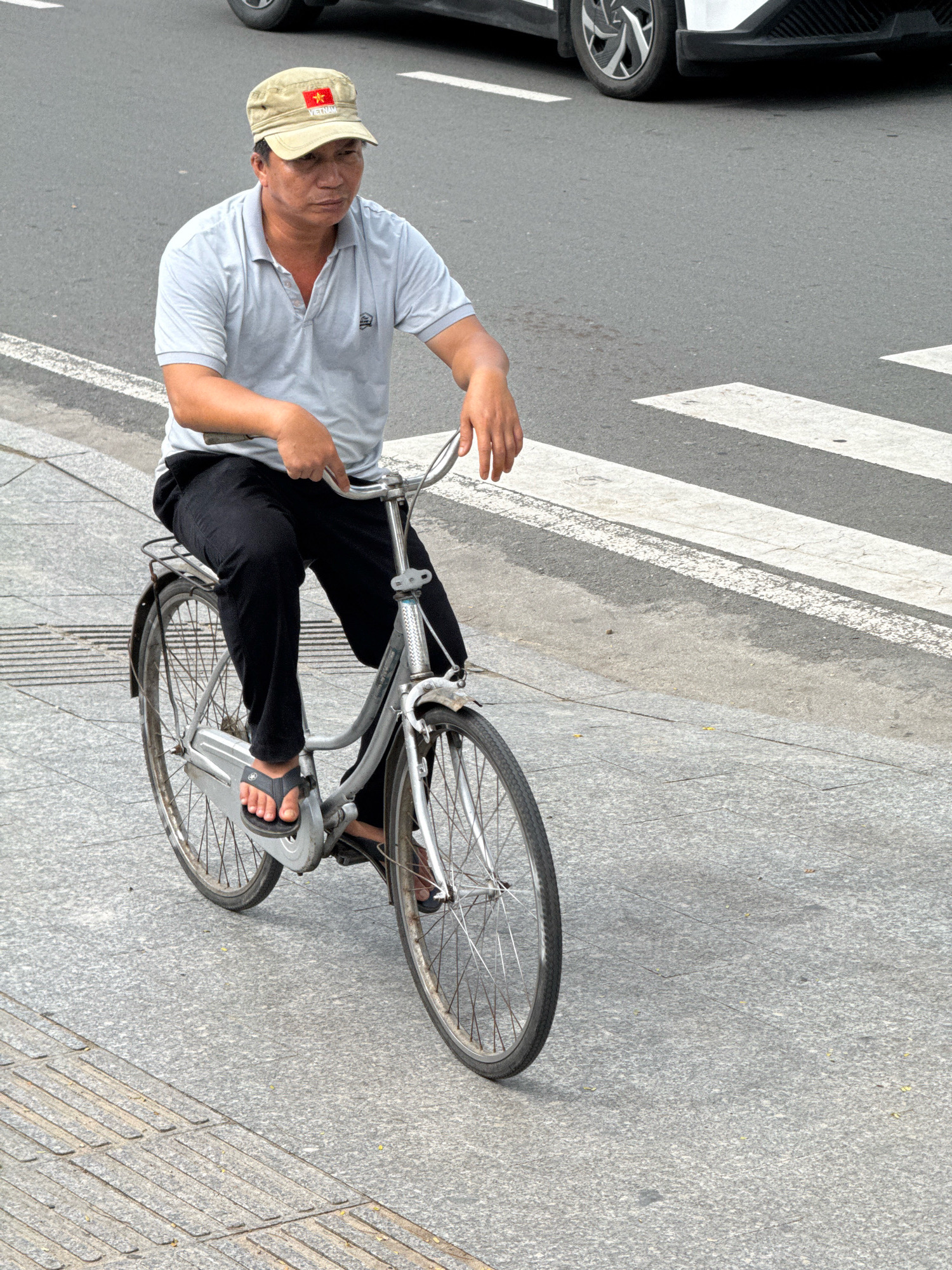 A middle-aged man wearing a beige cap with a Vietnamese flag, a light gray polo shirt, and black pants is riding a silver bicycle on a paved street in Nha Trang, Vietnam. The man is looking to his right. The bicycle has a rear rack. The street has a white pedestrian crossing marked on it. A white car is partially visible in the background. The ground is a light gray speckled pavement.