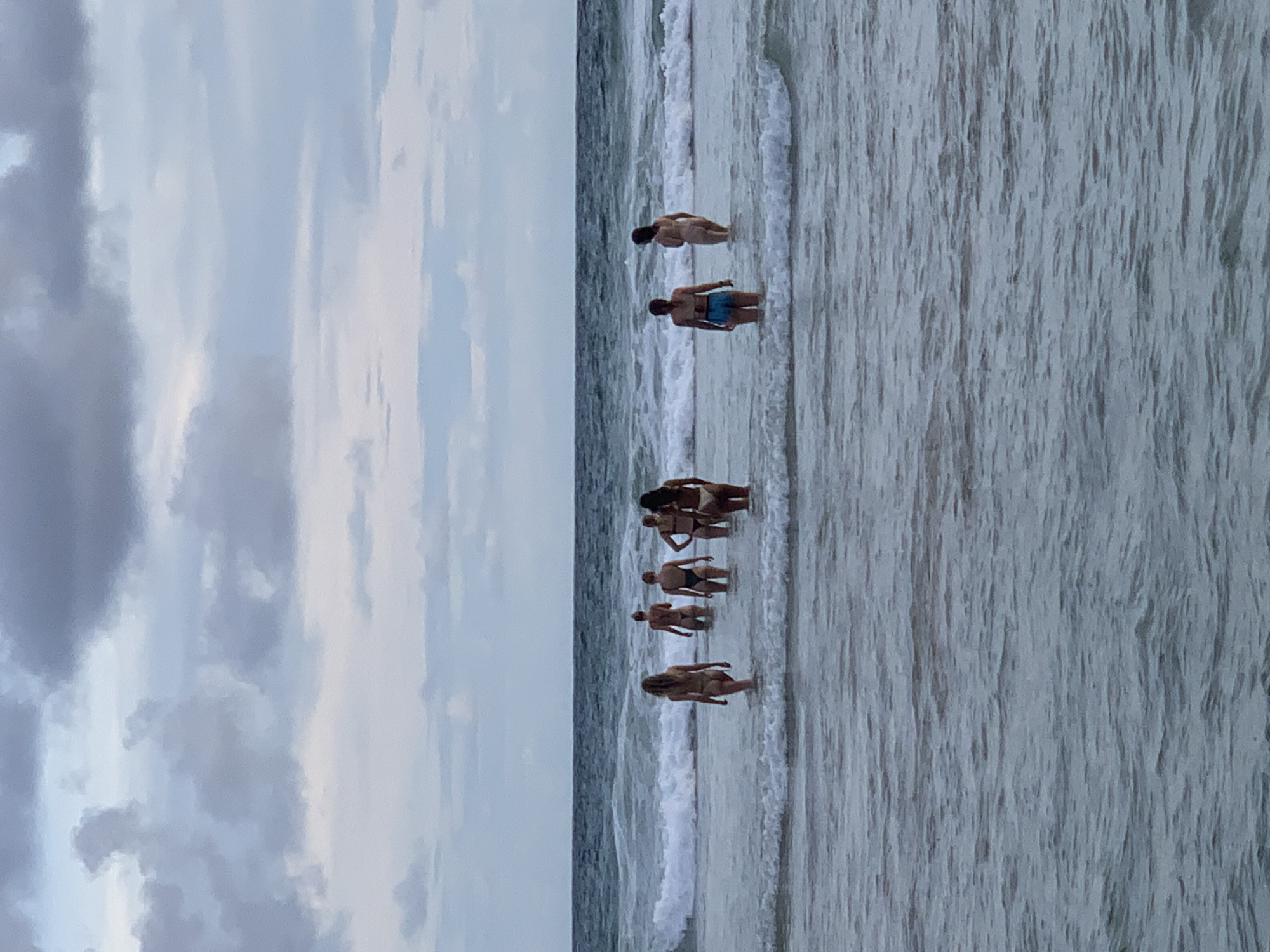 People stand in the ocean on the coast of Kathaluwa, Sri Lanka, under a cloudy sky. A group of seven people is wading in the water, and two more people are farther out from the group. The ocean appears turbulent with white foam.