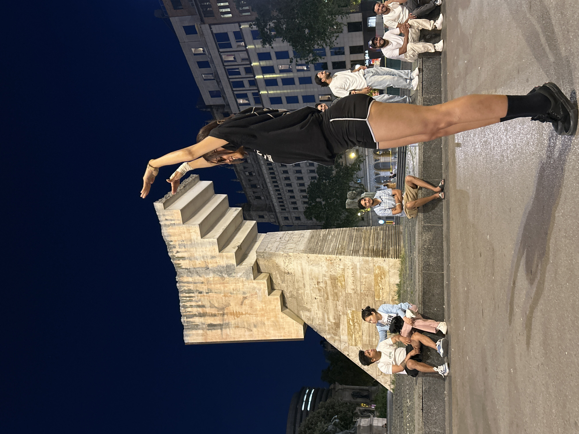 Here is a detailed description of the image:
The image shows a scene in Barcelona, Spain at night, centered around the Cascade Monument, located in the Ciutadella Park. The sky is dark blue. A group of people is sitting or standing on the pavement, with the Cascade Monument providing a unique architectural backdrop.
One individual, appearing to be a young woman, is posing in a horizontal, plank-like position near the steps of the Cascade Monument, using it as a prop. She is dressed in a dark t-shirt and black shorts, with black socks and shoes. Her arms are extended towards the monument.
Scattered around her are other individuals, likely a group of friends or onlookers. Some are seated on the ground, while others stand or sit on the elevated platform of the monument. In the background, a building with multiple illuminated windows is visible. The ground appears to be wet.
