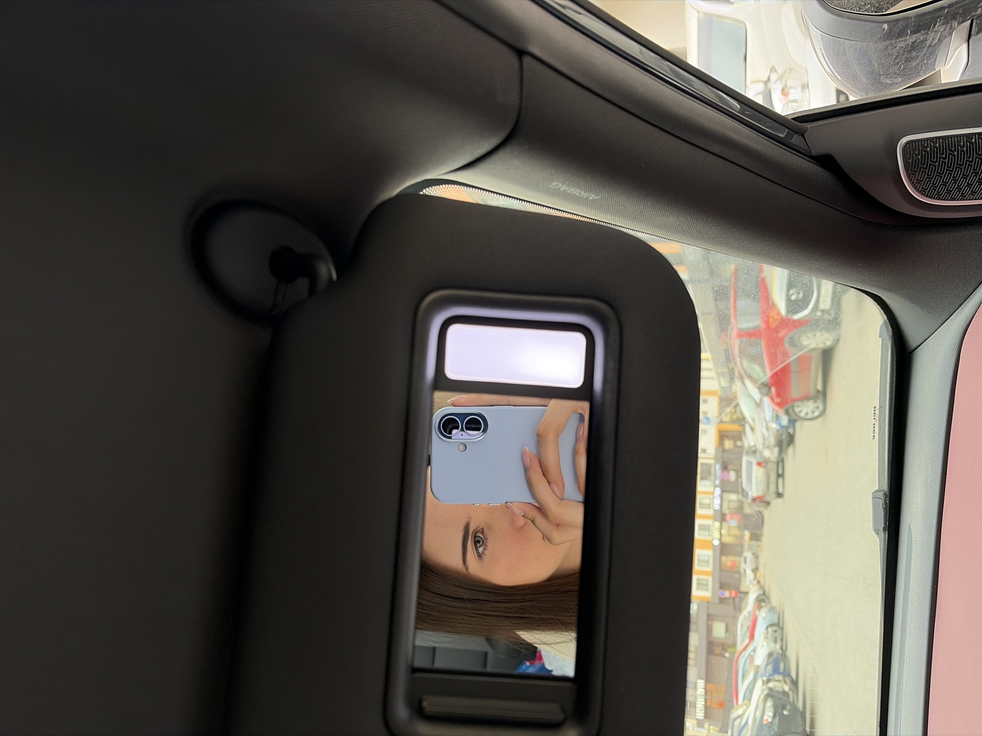 The image shows the interior of a car, specifically the driver's side sun visor. A woman is taking a selfie in the visor's mirror, holding a light blue phone. Through the car window, a street in what appears to be a parking lot is visible, lined with parked cars and buildings. According to a 'BERGEE' sticker visible on the edge of the car window, the location is Izhevsk, Russia.