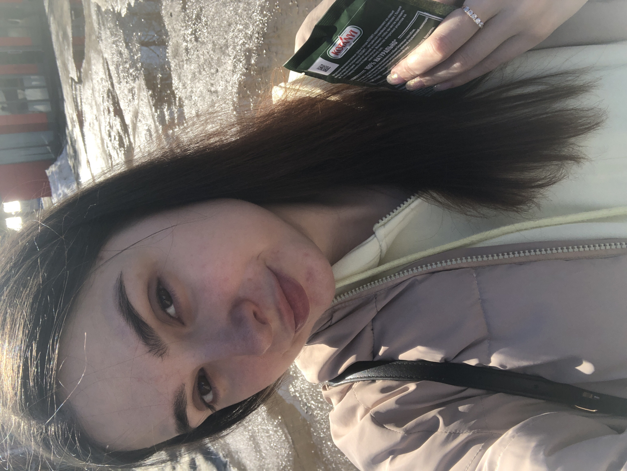 Here is a detailed description of the image:
The image is a selfie of a young woman with dark hair, fair skin, and prominent eyebrows. She is holding a packet of "KOTANYI" spices in her right hand, displaying it towards the camera. She is wearing a ring on her ring finger.
She is wearing a light-colored, possibly beige, padded jacket over a yellow hoodie. A black strap, possibly from a bag, is visible across her torso.
The background is somewhat blurred but appears to be an outdoor environment, possibly a city setting. The background appears to contain reflective surfaces, possibly ice or wet pavement.
The OCR text from the image indicates that "KOTANYI" is an Austrian family company with a range of herbs and spices. Also, it is mentioned that it is 100% natural.
Without additional context, I cannot confirm the exact location as Noviy, Russia; however, the text is in Russian, which suggests that the image was taken in a Russian-speaking country.