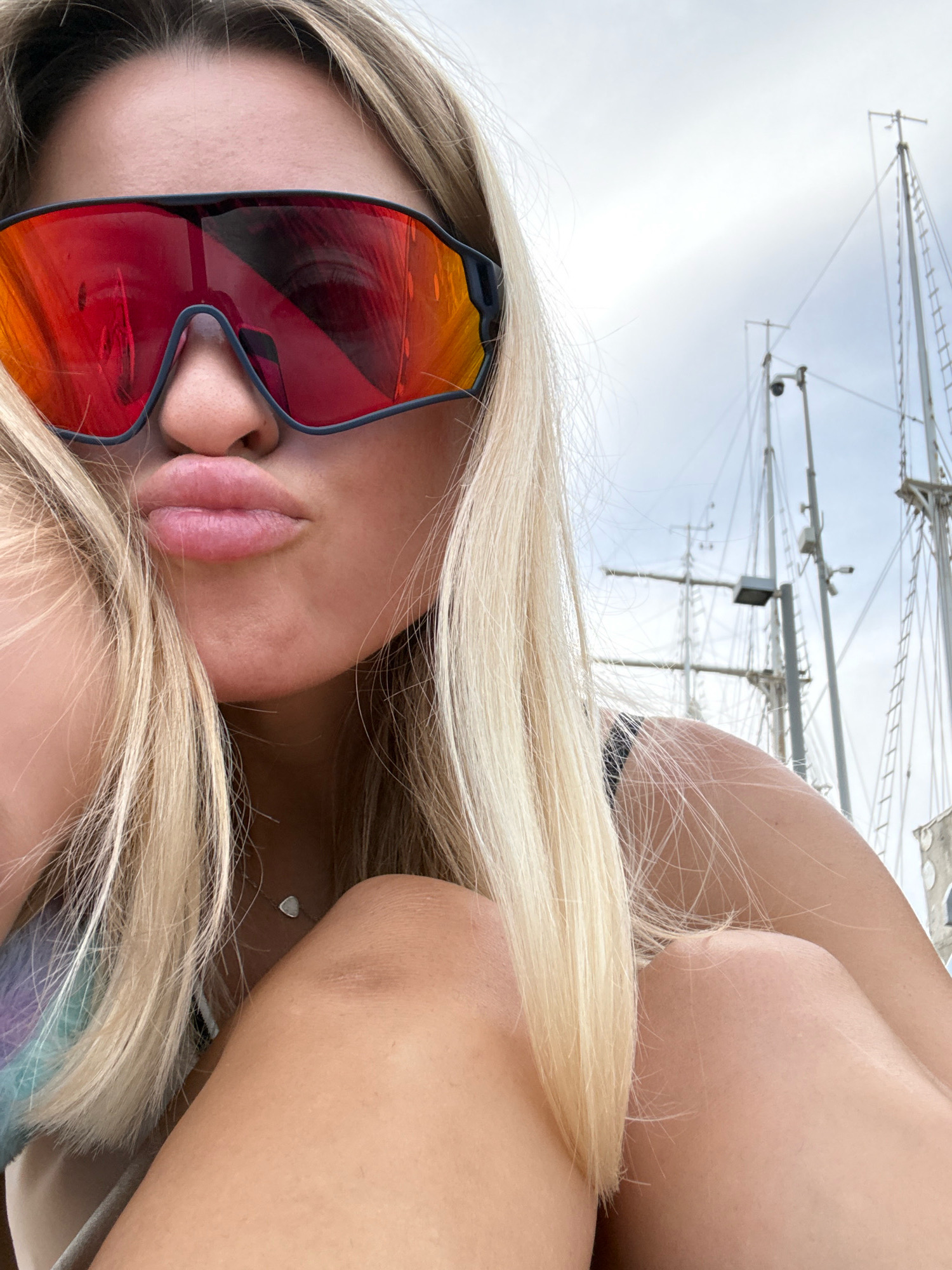A close-up, waist-up shot shows a fair-skinned woman with blonde hair wearing large, red-tinted sunglasses. She is puckering her lips. Her hair is parted to the side and falls around her face and shoulders. A hint of a purple and blue patterned garment is visible on her left side. She is wearing a delicate silver necklace with a small, light-colored pendant. Her right arm is bent, resting on her lap. In the background, multiple tall ship masts with rigging are visible against a cloudy sky, suggesting a location near a harbor or marina. The image appears to have been taken outdoors, possibly in Valencia, Spain, given the presence of sailing vessels.