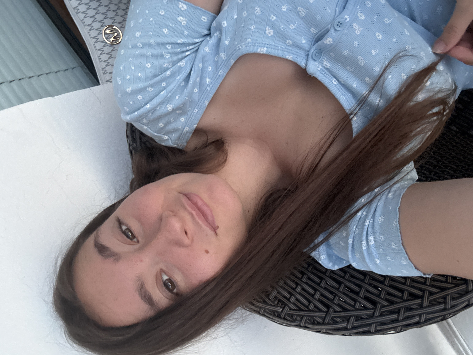A young woman is captured in a close-up, overhead shot, lying on a wicker chair. Her head is tilted back, and she looks directly into the camera with a relaxed and serene expression. Her long, dark brown hair falls around her face and shoulders. She is wearing a light blue, short-sleeved top with a delicate white floral pattern. A single button is visible at the neckline of her top. To her left, a white surface with a repeating "M" pattern and a circular gold emblem with a stylized "Z" are visible. The background is blurred, but suggests an outdoor setting with natural light. The overall mood is casual and peaceful. No text is clearly legible in the image.