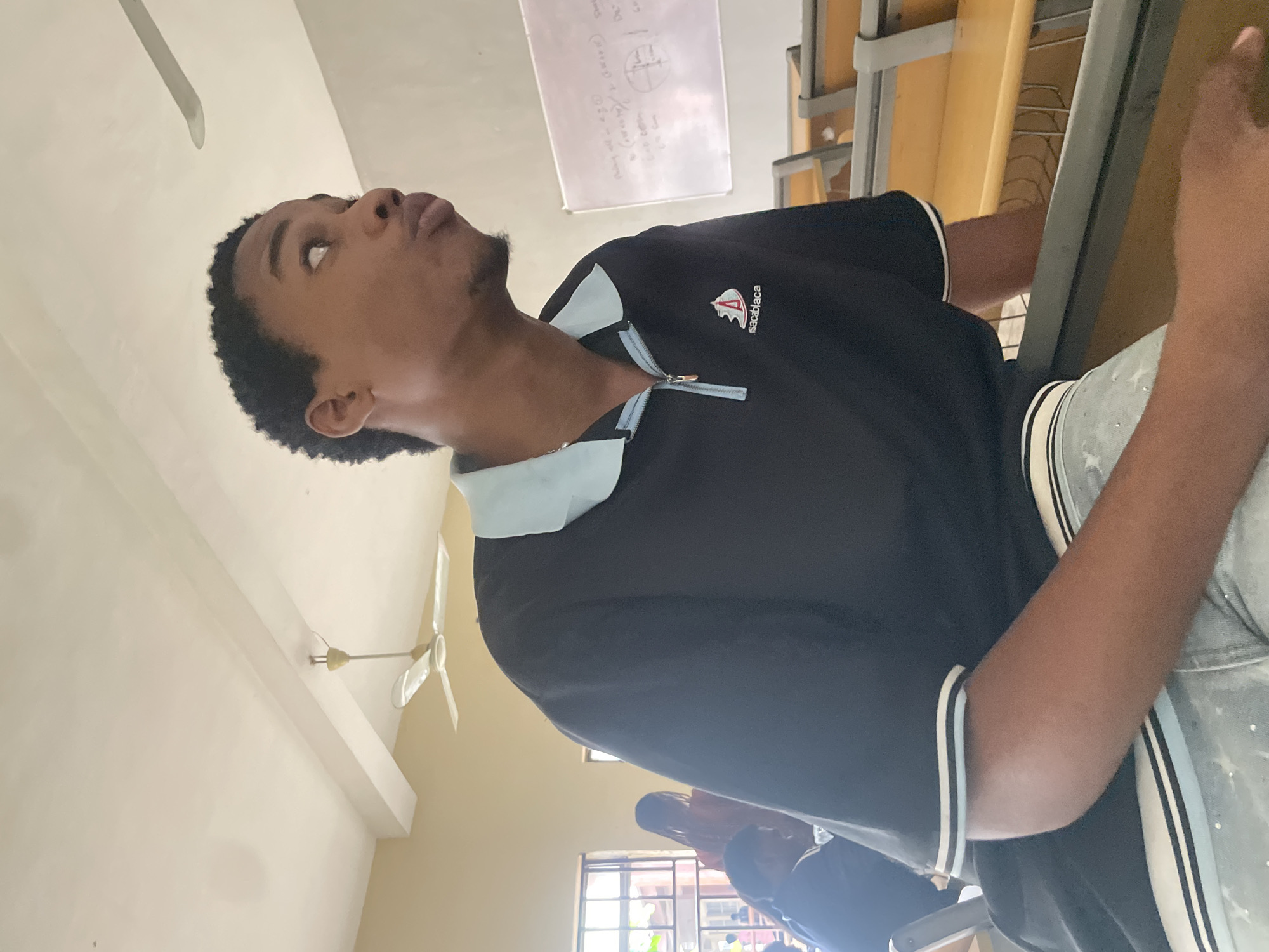 In Ihiagwa, Nigeria, a young man with dark skin and short, curly hair is captured in a medium shot, looking to his right with his lips puckered. He is wearing a black polo shirt with a light blue collar and a zippered neckline, featuring a small logo with a boat and text that appears to read "sacablaglaca". His light-colored, speckled jeans are also visible. He is seated at a desk in what appears to be a classroom or lecture hall, with rows of yellow desks and chairs stretching behind him. Natural light streams in from a window on the left side of the frame, illuminating the scene and casting soft shadows. A white ceiling fan is visible in the upper left portion of the image, and a whiteboard with some writing can be seen on the right wall. Other individuals are faintly visible in the background, seated at desks.