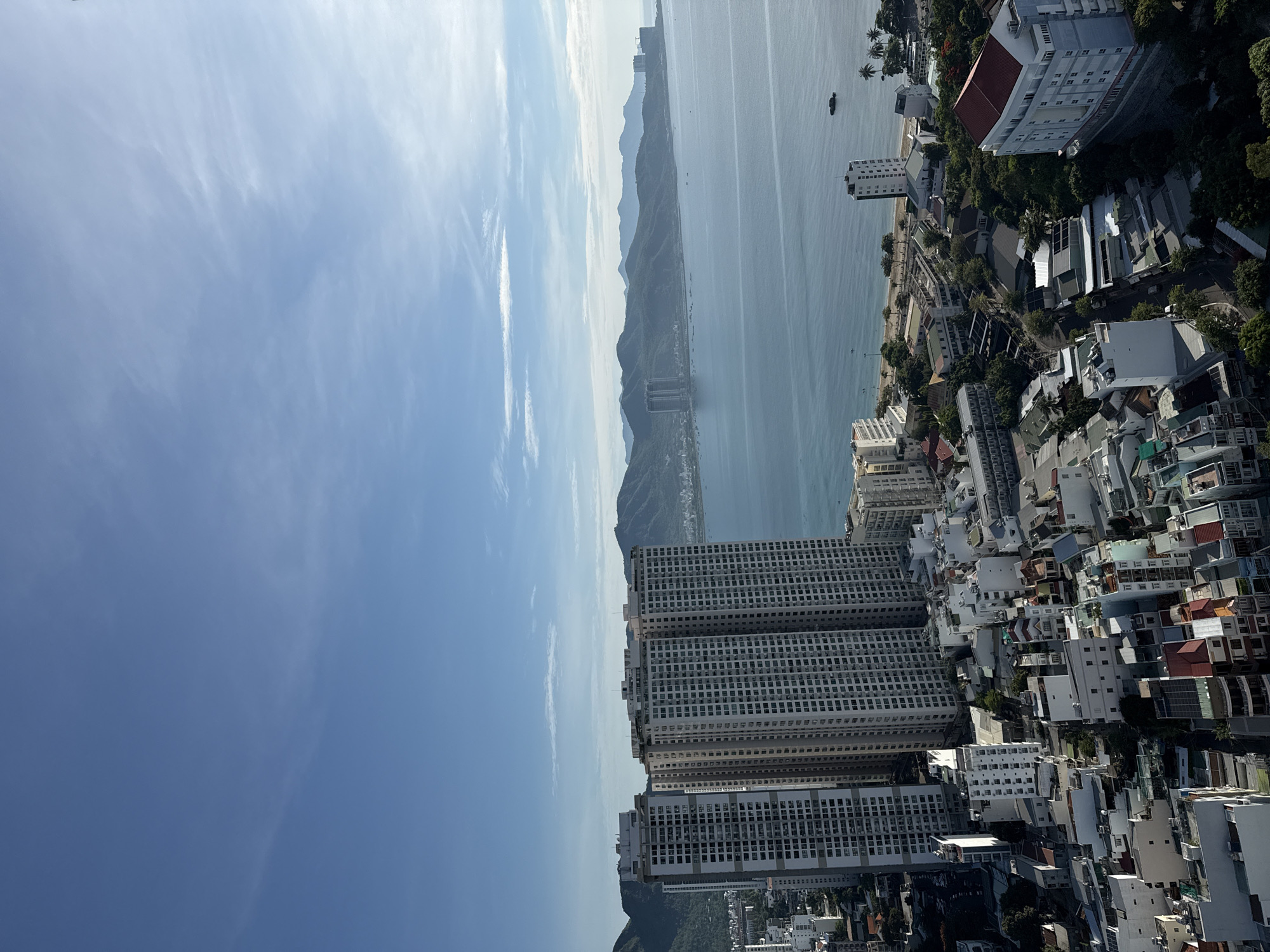 This image shows a panoramic view of the city of Nha Trang, Vietnam, from a high vantage point. The scene is dominated by a clear blue sky with wisps of white clouds. In the foreground, several tall, modern apartment buildings with numerous windows are visible. To the right of these buildings, a beach can be seen bordering a calm, blue ocean. The ocean stretches towards a distant, verdant mountain range that forms a natural backdrop to the city. Scattered along the coastline are various other buildings, including lower-rise structures and what appears to be a hotel with a red roof. The overall impression is a vibrant coastal city with a blend of high-rise architecture and natural scenery. There are no visible people or specific actions captured in this static view.