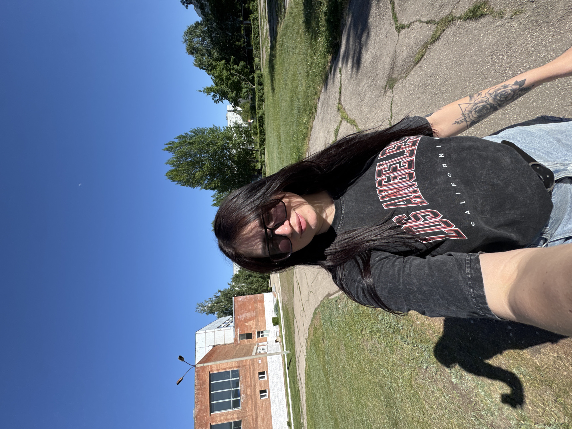 A selfie shows a woman with long, dark hair and sunglasses, wearing a black "Los Angeles California" t-shirt. She stands outside on a sunny day with a bright blue sky above. The background includes a green lawn, a cracked concrete pathway, and a brick building with white window frames. Trees are visible in the distance. The woman has a tattoo on her left arm. Her shadow is cast on the grass. The image is rotated counter-clockwise. Location: Unknown, Unknown.
