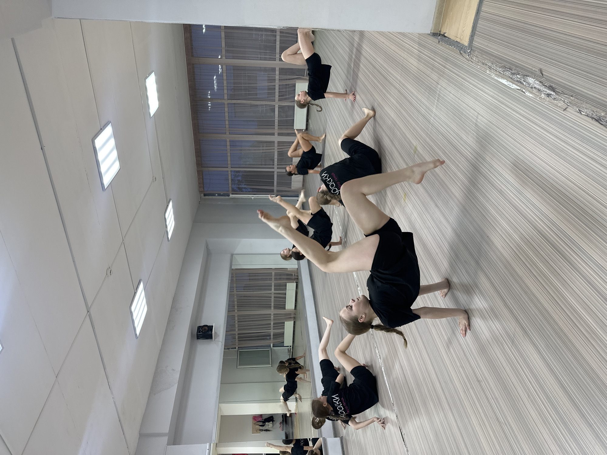 A group of at least seven young people are in a dance studio in Votkinsk, Russia, practicing yoga or gymnastics-type movements. They are all dressed in black shirts and shorts. The room has light-colored walls and wood-grained flooring, with a large mirror along one wall. Rectangular lights are recessed in the ceiling. Outside the windows, it appears to be dusk or evening.