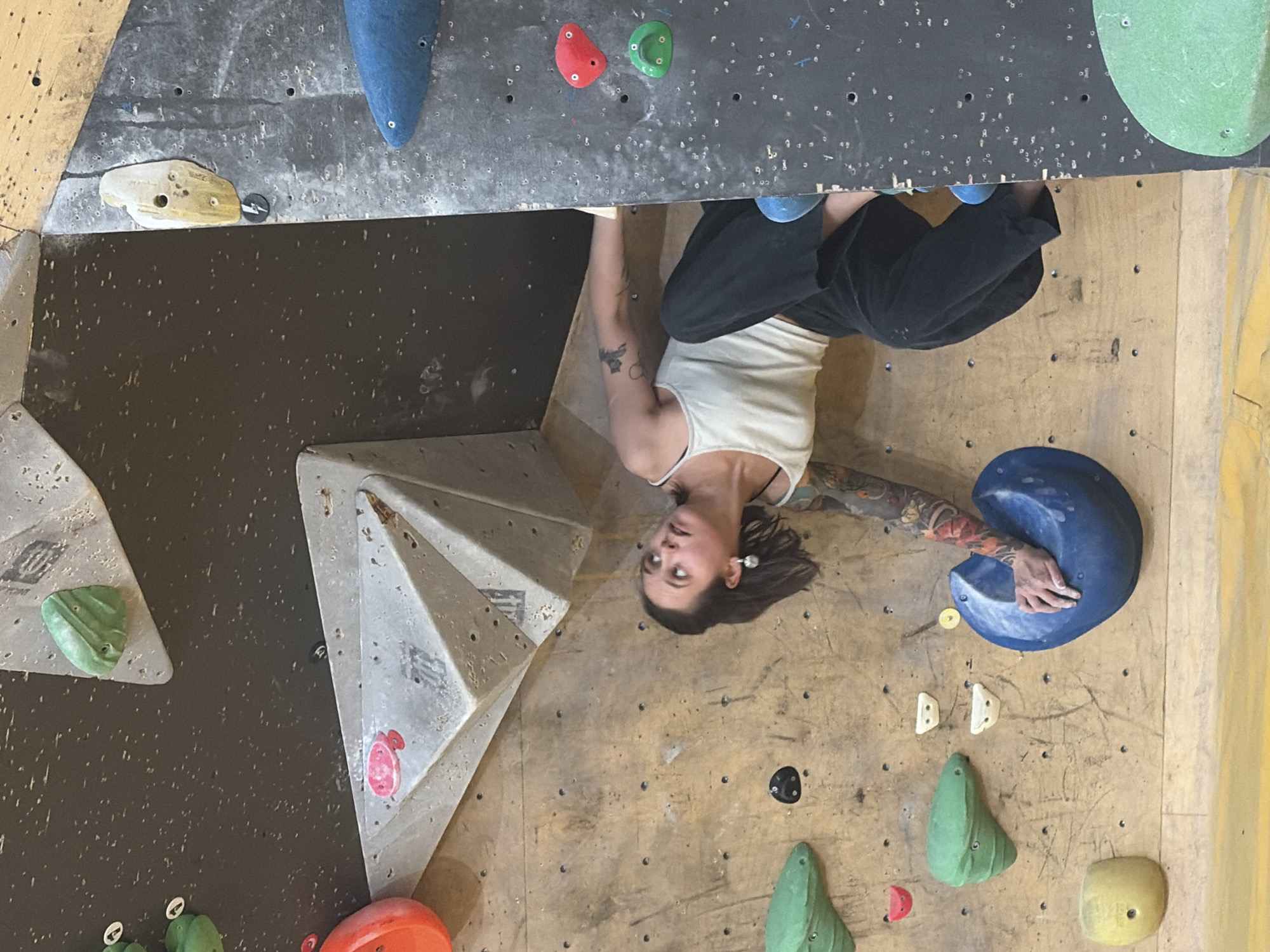 The image shows a woman with dark hair, wearing a white tank top and black pants, climbing a bouldering wall. She has tattoos on her arms. She is gripping a blue climbing hold and looking upwards. The bouldering wall has various colored climbing holds, including green, red, yellow, and blue. The wall is made of a textured material, with some sections appearing to be wood and others a darker, speckled surface. There are several other climbing holds visible on the wall, as well as wooden paneling at the bottom of the frame. The lighting suggests an indoor setting. The image was taken in Moscow, Russia.