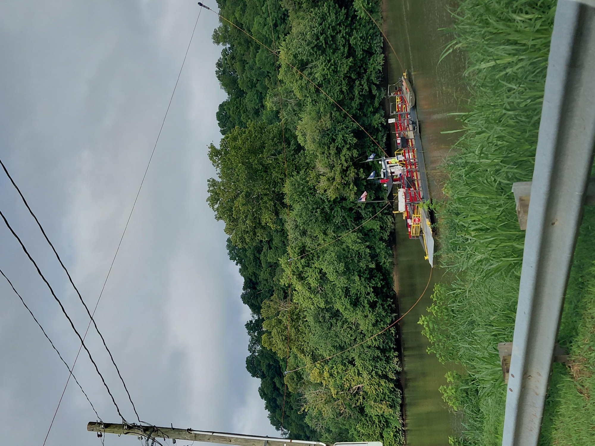 An outdoor, eye-level, medium shot captures a ferry crossing a river on a cloudy day. A wooden utility pole with several wires extends from the left edge of the frame towards the top. The ferry, a flat barge with a small cabin structure, is positioned in the center of the river, connected to cables that run to the shore on either side. The ferry has red railings and appears to be carrying at least one person wearing a yellow safety vest inside the cabin. Two flags, one American and one blue, are visible on the ferry. The riverbanks are lined with lush green trees and vegetation. In the foreground, a silver guardrail separates the viewer from a grassy embankment. The water is calm and reflects the overcast sky and surrounding trees. The scene suggests a rural or semi-rural setting, likely a crossing point for vehicles or pedestrians. This image is not in the city of Richmond, United States; it depicts a river crossing in a more natural, wooded environment.