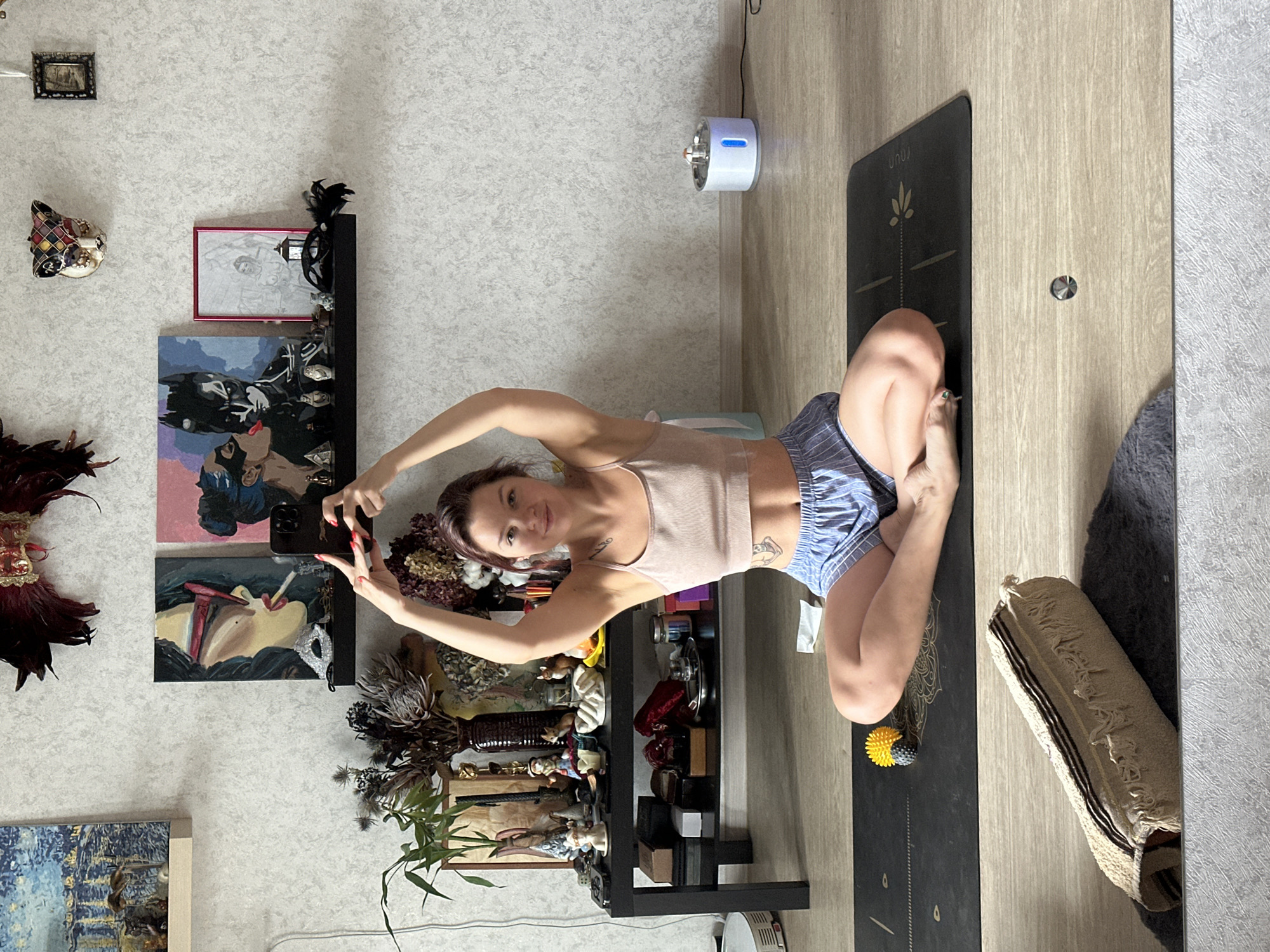 A young woman with auburn hair is sitting cross-legged on a yoga mat, holding her hands above her head. She is wearing a light pink tank top and blue striped shorts. The room appears to be a living area, with a shelf displaying various decorative items and paintings on the wall. A black coffee table is visible to the left, with plants and other objects on it. A cat water fountain is on the floor to the right. The woman is holding a smartphone in her right hand, taking a selfie. The floor is made of light-colored wood. The overall scene depicts a domestic setting. There is no identifiable information about the city of Vladimir, Russia.