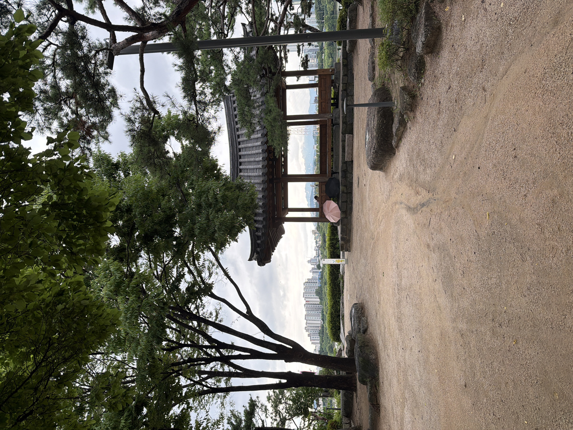 The image depicts a tranquil scene in a park in Seoul, South Korea. In the foreground, a dirt path winds through a garden area with large rocks and lush green trees. A traditional Korean gazebo, with a dark tiled roof and wooden pillars, stands in the middle ground. Two people are seated inside the gazebo, one holding a pink umbrella. In the background, a cityscape with numerous apartment buildings is visible, suggesting the park is situated within a densely populated urban area. The sky is overcast, with a hint of distant mountains. The overall atmosphere is peaceful and serene.