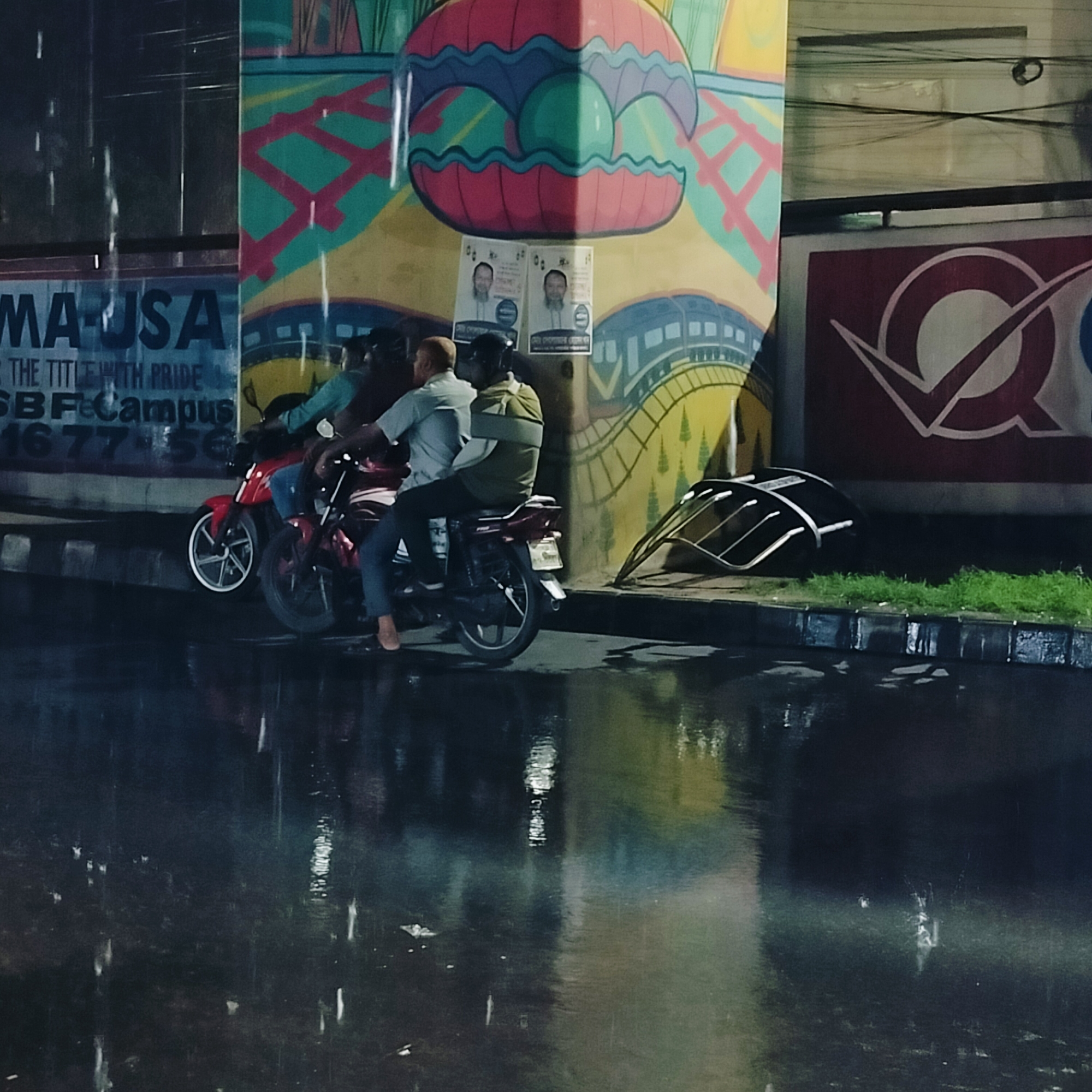 The image captures a rainy street scene in Dhaka, Bangladesh, at night. Two motorcycles are parked near a pillar adorned with colorful graffiti and posters. The motorcycles are occupied by three men, one on the first and two on the second, one wearing a helmet. The street surface is wet, reflecting the surrounding lights and rain. In the background, signs and logos are visible on nearby buildings. The overall atmosphere suggests a typical urban scene during a rain shower.