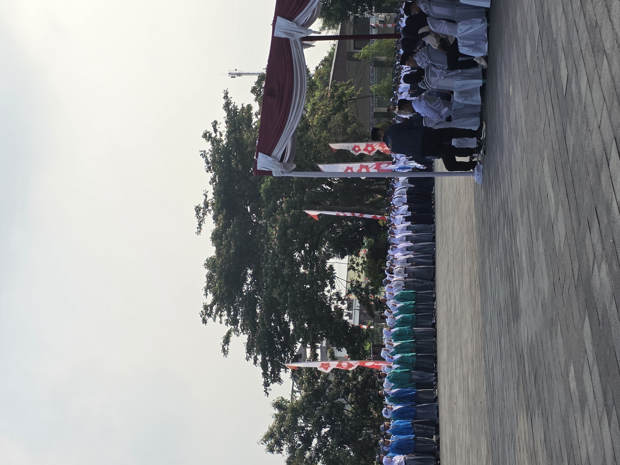 This vertical, eye-level, outdoor photograph depicts a formal gathering of students and adults in what appears to be Tambun, Indonesia, given the context. The scene is set in an open, paved area, likely a school or public plaza, with a light gray sky overhead.
In the foreground, a dark gray paved ground is visible, with a pattern of rectangular stones. To the right, a white tent with a maroon trim is set up, with several chairs draped in white fabric placed beneath it. A man in a dark jacket and black pants is standing near the tent, facing away from the camera.
Further back, a large group of students is arranged in neat rows, facing left. The majority are wearing school uniforms: white shirts with dark skirts or pants, and either blue or green jackets or vests. Some of the female students are wearing hijabs. A few adults, possibly teachers or officials, are also present among or behind the students.
Several tall, thin flags with red and white patterns are planted in the ground among the students, indicating a possible national or school event. Large, mature trees with dense green foliage are visible in the middle ground, behind the students. In the distance, buildings and a cellular tower can be seen against the hazy sky. The overall impression is one of an organized assembly or ceremony.