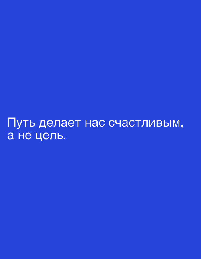 The image is a close-up of a blue background with white text. The text reads: "Путь делает нас счастливым, а не цель." This translates to "The journey makes us happy, not the goal." There are no people, subjects, objects, actions, interactions, or environmental details visible in the image. The provided location information is not relevant to the image content.