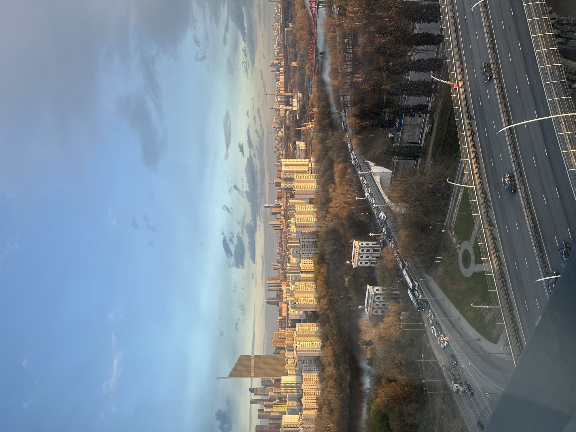 A high-angle, outdoor shot captures a cityscape. The sky, a gradient of pale blue to gray, occupies the left third of the frame. Below the sky is a dense urban area with buildings of varying heights, arranged in linear patterns along roads. A river runs through the middle of the city and trees line the river. The highway with vehicles is present on the right side of the image. A red bridge is also seen further back in the distance. This is most likely a view of the city of Khimki, Russia.