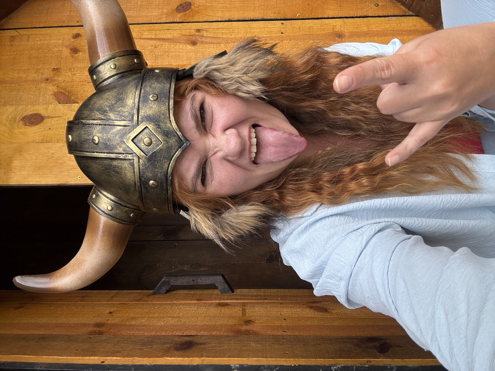 A woman with long, wavy red hair, wearing a golden Viking helmet with large brown horns and brown fur trim, sticks her tongue out at the camera. She is wearing a light blue, long-sleeved shirt. Her right hand is raised, forming a devil horns gesture. The background consists of a wooden structure with vertical planks of varying shades of orange and brown. A dark, recessed doorway is visible behind the woman's head. The scene appears to be outdoors, possibly at a tourist attraction or event, given the costume. There is no discernible indication of the location being in Thôn 1, Vietnam.