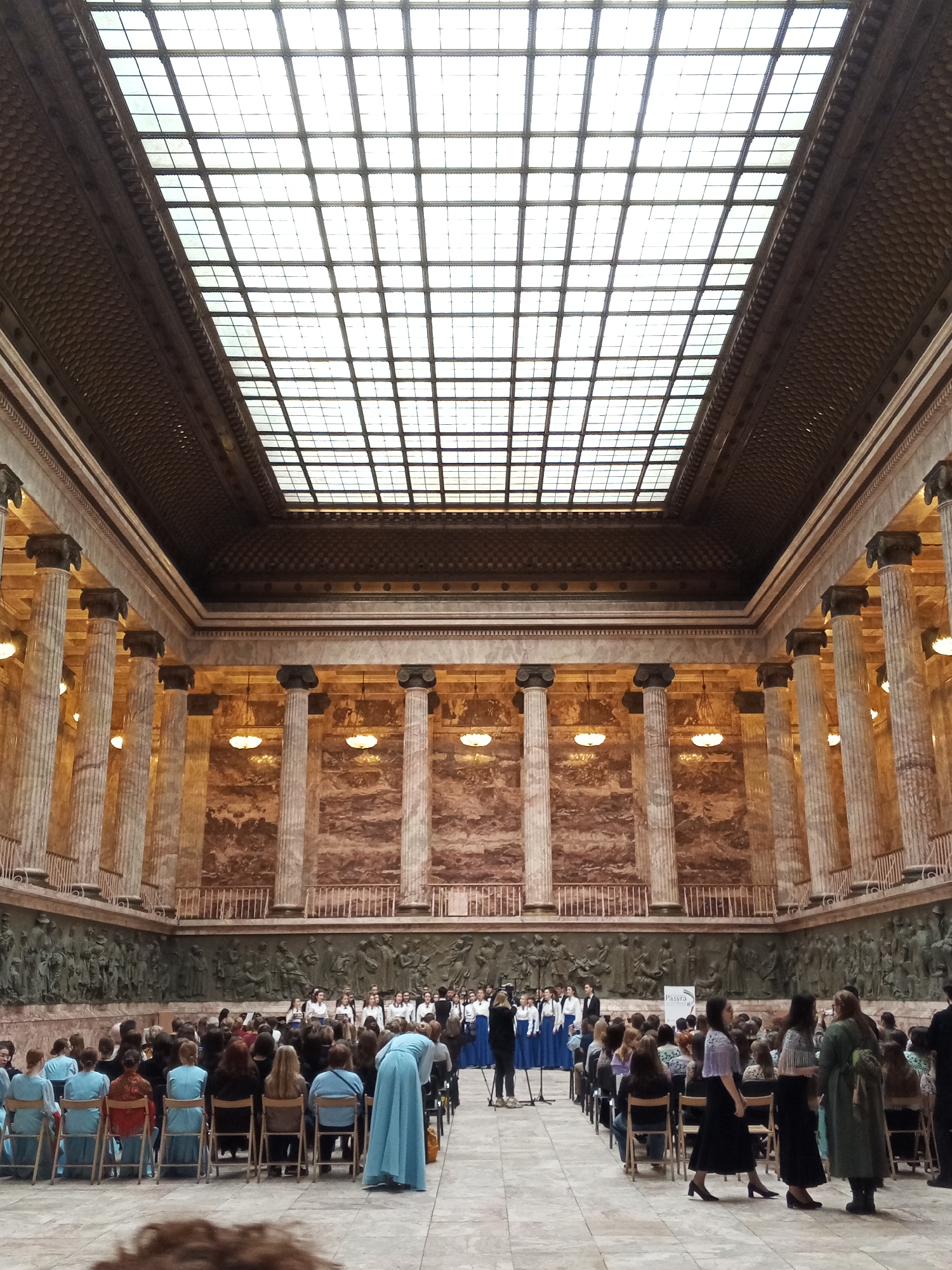 An interior scene, Saint Petersburg, Russia. A large room with a high ceiling featuring a gridded skylight is populated by a seated audience facing a choir standing center. Tall Corinthian columns line the walls and the walls have bas-relief sculptures. A woman in a blue gown is in the lower-center foreground. Several people are standing on the right. Many of the audience members are seated in rows of chairs. The overall ambiance is formal and subdued.