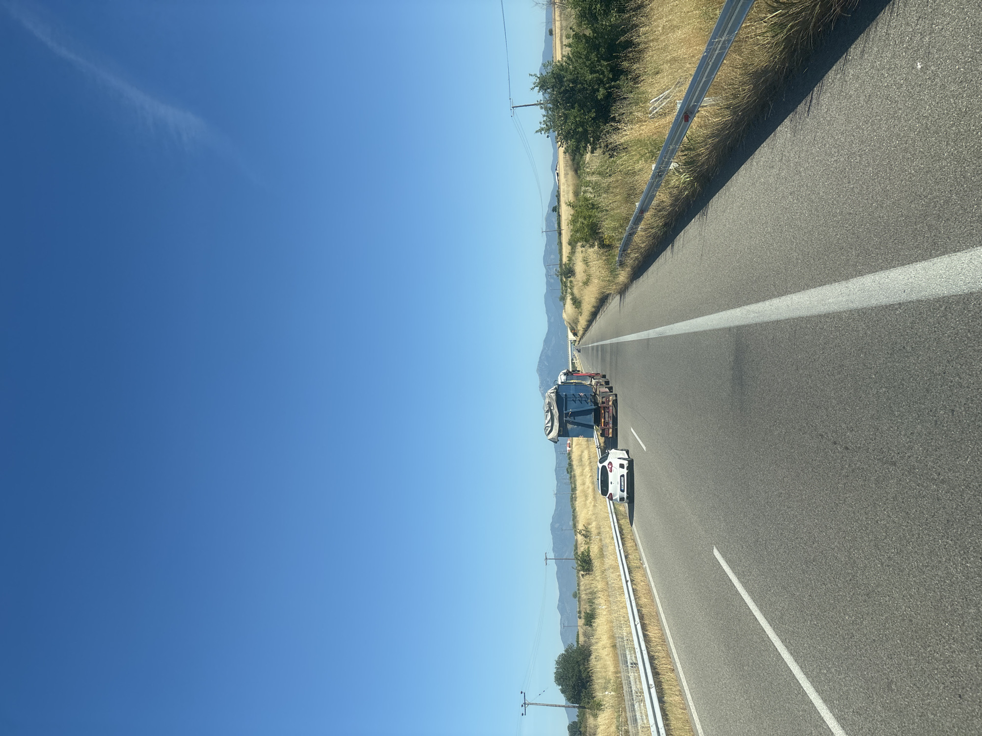 A bright, clear blue sky fills the upper half of the image, with a wispy white cloud stretching across the right side. The lower half depicts a two-lane asphalt road running through a rural, dry landscape under bright sunlight. The road is divided by a solid white line. On the left lane, a white car is traveling behind a large, blue flatbed truck carrying a covered load. The truck has a red cabin and is moving away from the viewer. Guardrails with metal posts and reflective markers line both sides of the road. The terrain on either side is covered in dry, tall, yellowish-brown grass, with scattered green bushes and trees further in the distance. Several utility poles with wires are visible along the roadside, and a range of hazy mountains can be seen on the horizon under the clear sky. The overall scene suggests a highway or major road in a dry, Mediterranean climate, consistent with the region of Kallithea, Greece, during a sunny day.