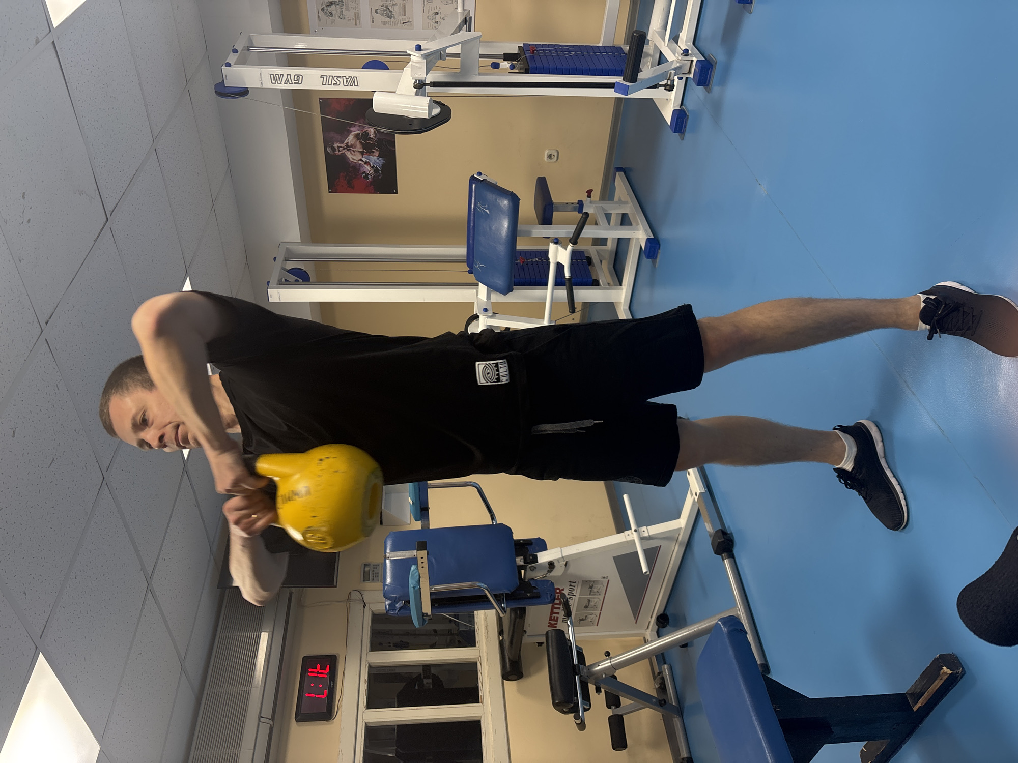 The image shows a man in a gym holding a yellow kettlebell. He is wearing a black t-shirt and black shorts. The gym has blue floors and various weight training machines. A poster is on the wall, and there is a digital clock displaying a time. The gym is identified as "VASIL GYM." The location is Chaikovsky, Russia.